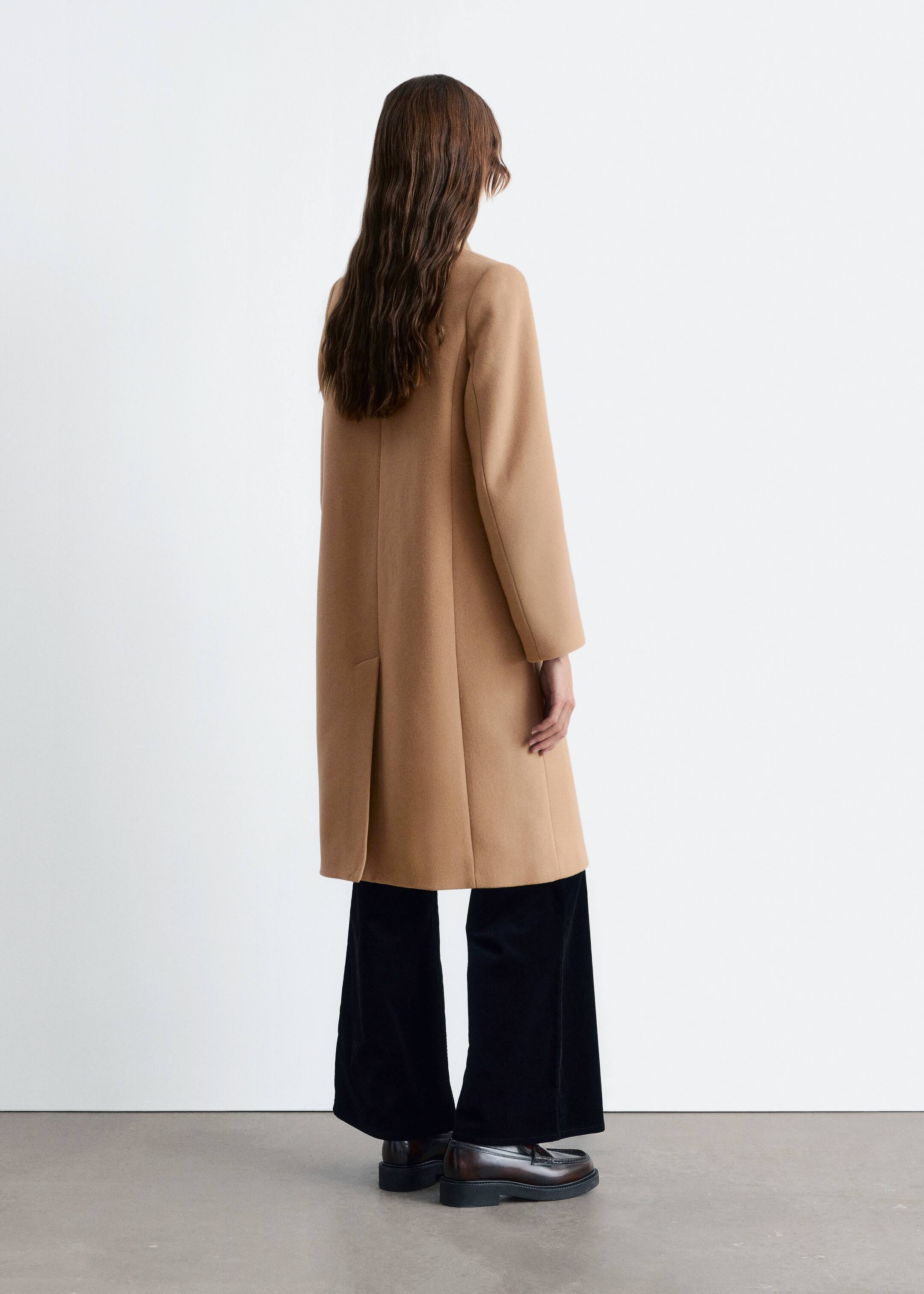 View larger image: A camel-colored long coat is being viewed from the back, showcasing its straight, relaxed silhouette. It is featuring a single vent at the lower back and is falling to a mid-calf length, layering over dark wide-leg trousers and dark loafers.