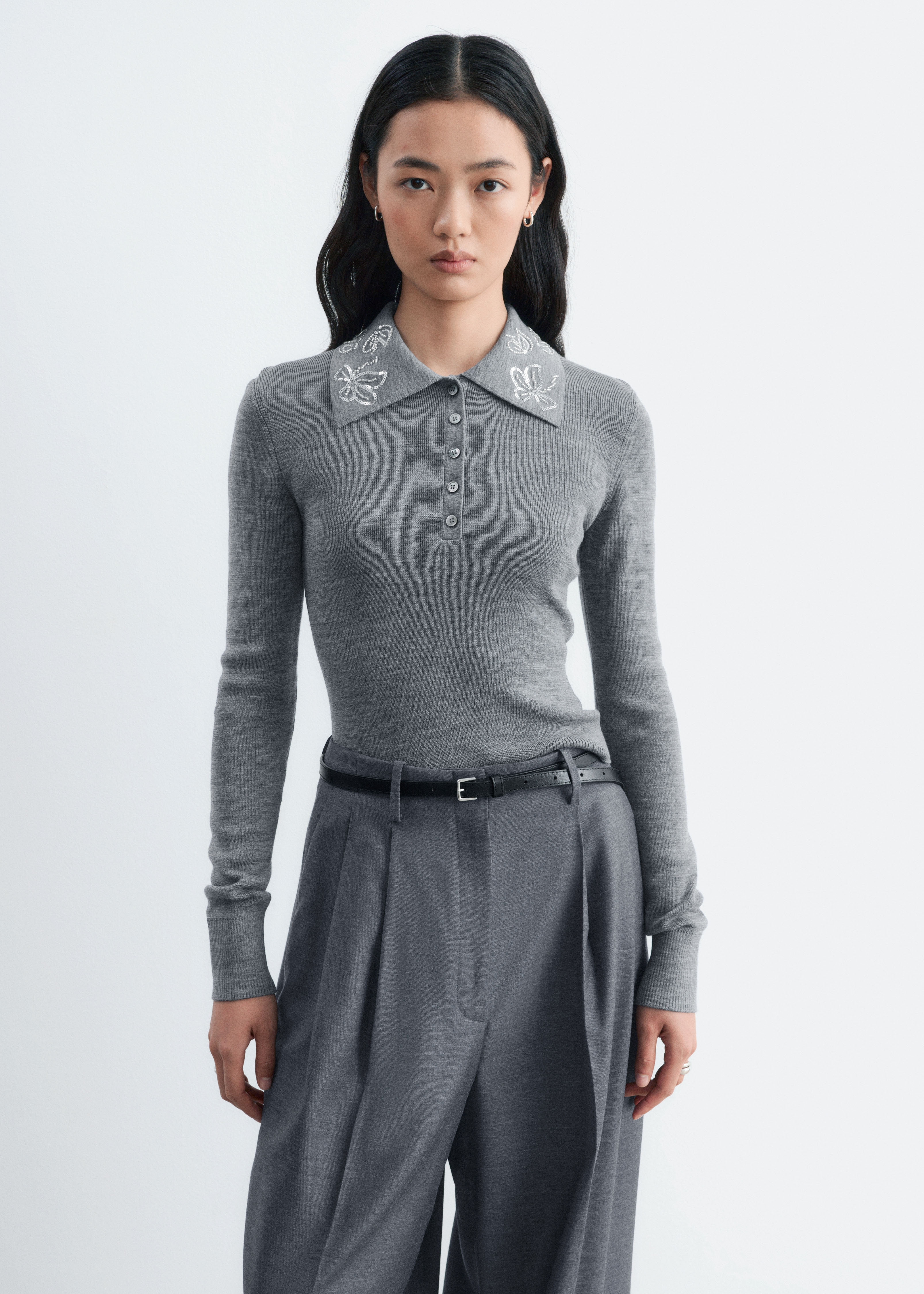 Un maglione polo in lana grigia mostra una vestibilità slim-fit sul davanti, mettendo in risalto un colletto a punta impreziosito da un ricamo a farfalla con paillettes argentate. Il maglione presenta una chiusura a cinque bottoni sul petto e maniche lunghe con polsini a coste.