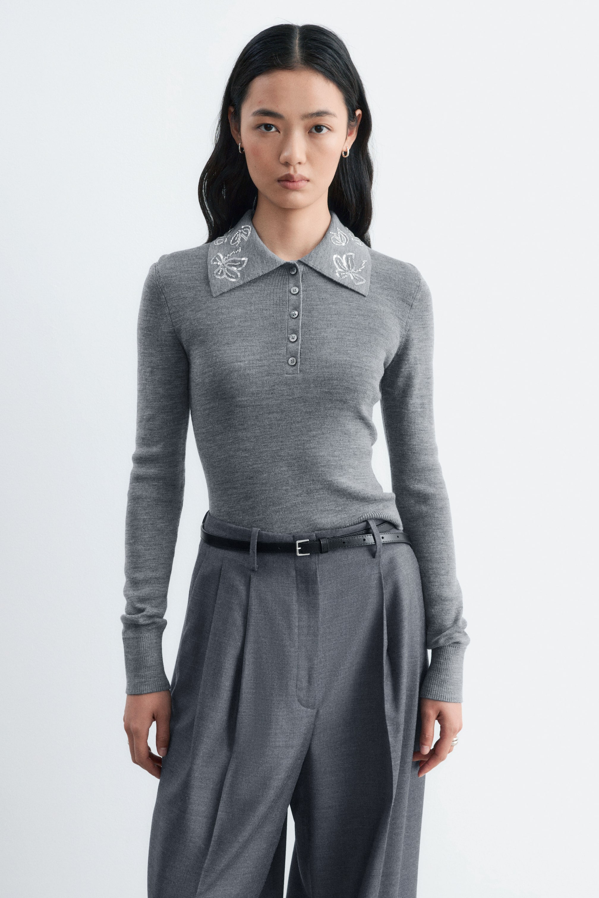 Agrandir l'image: Un pull polo en laine gris à manches longues, ajusté et doté d’une patte de boutonnage, est porté par le mannequin. Le pull présente une broderie florale argentée sur le col et est rentré dans un pantalon gris à jambes larges, maintenu par une fine ceinture noire.