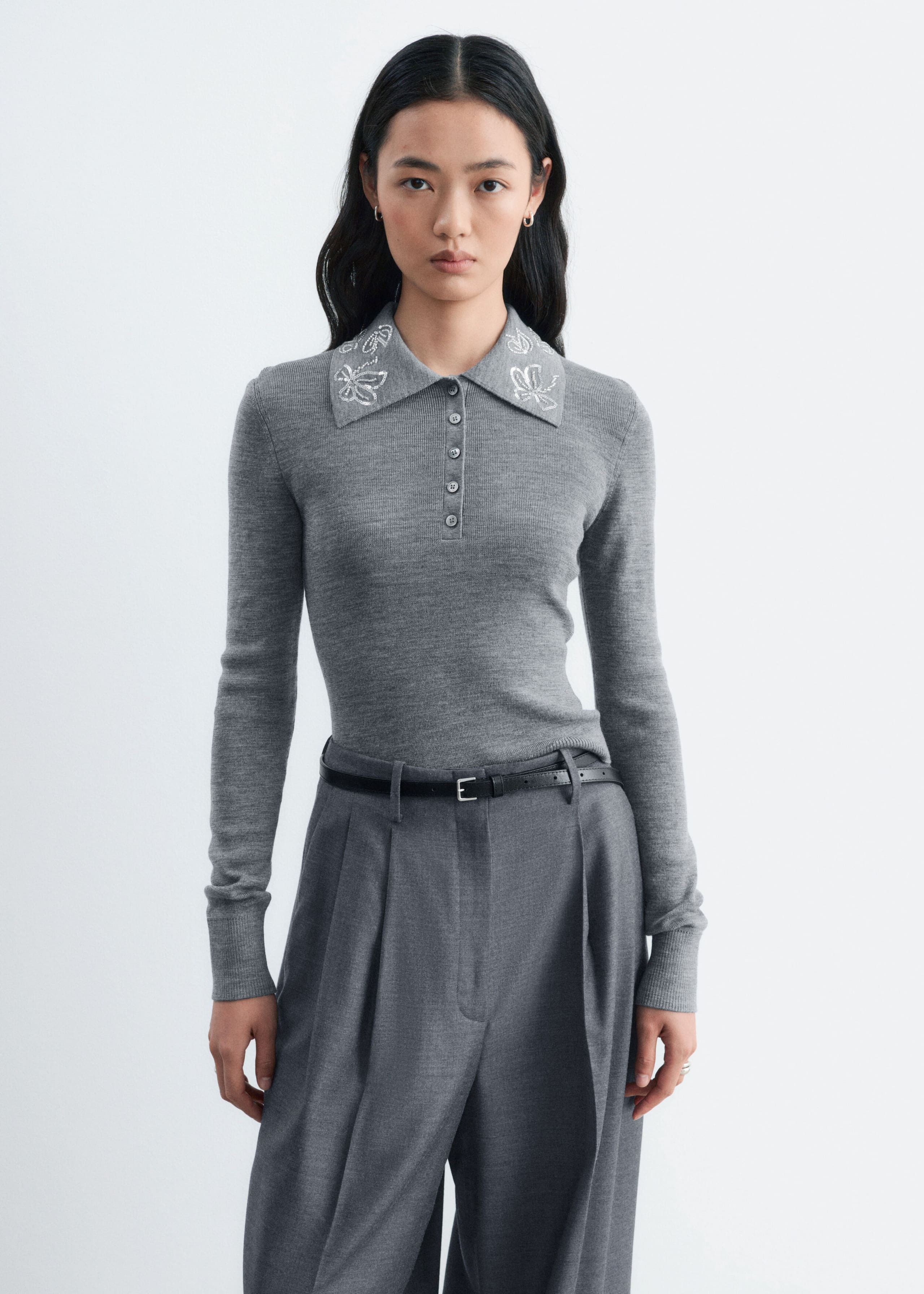 Ein grauer Woll-Pullover mit langen Ärmeln, schmaler Passform und Knopfleiste wird vom Model getragen. Der Pullover zeigt silberne Blumenstickerei am Kragen und ist in eine weite graue Hose gesteckt, die mit einem dünnen schwarzen Gürtel gesichert ist.
