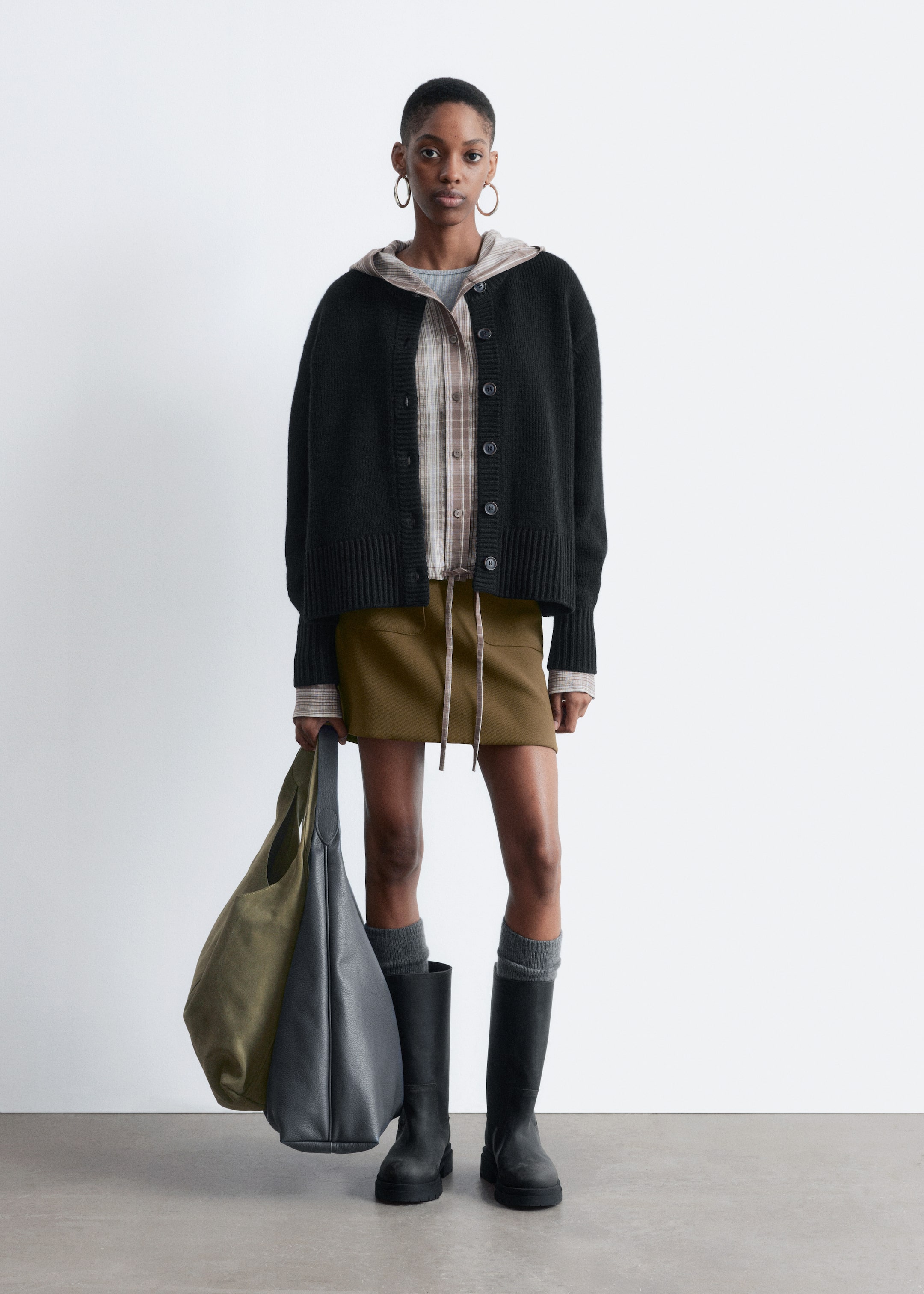 Agrandir l'image: Un cardigan en tricot noir surdimensionné avec un devant boutonné et des poignets et ourlet côtelés est porté superposé à une chemise à carreaux et un haut à capuche gris, associé à une mini-jupe vert olive dotée d'une taille à cordon. La tenue est complétée par des chaussettes grises jusqu'aux genoux, des bottes à semelles épaisses noires, et un sac cabas souple bicolore vert olive et gris foncé tenu, le tout présenté sur une silhouette debout faisant face à l'avant.
