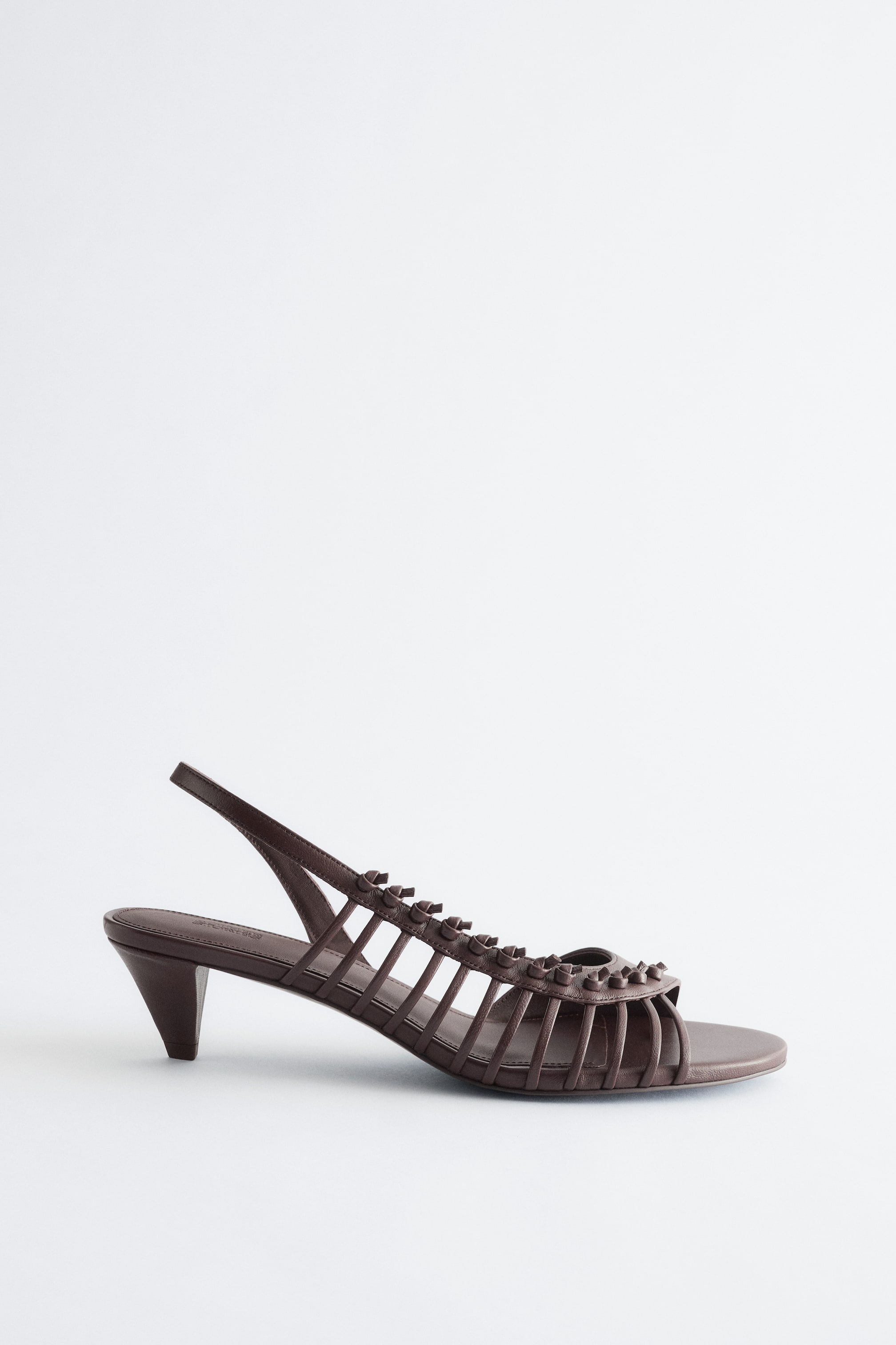 Zobacz większy obraz: Ciemnobrązowy sandał na pasku, skierowany w lewo, z wieloma paskami tworzącymi kratkowy wzór na przedniej części stopy, ozdobiony małymi ćwiekami i niskim stożkowym obcasem.