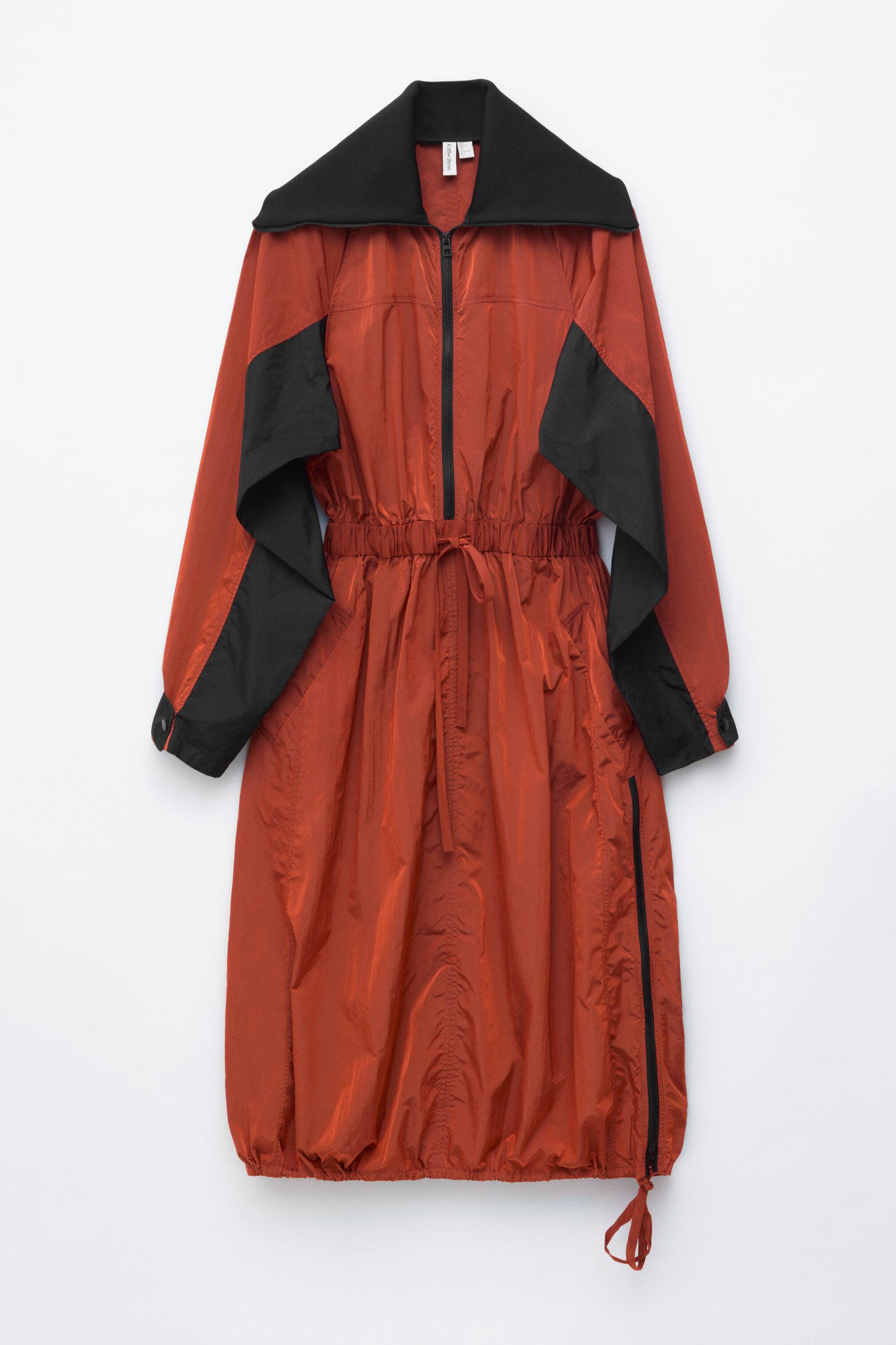 Agrandir l'image: Une robe midi est présentée par FRONT, confectionnée dans un tissu technique couleur rouille avec des empiècements noirs sur les épaules et les manches. Elle est dotée d'un col montant noir, d'une encolure zippée à l'avant, d'une taille à cordon de serrage créant des fronces, et d'un ourlet légèrement froncé avec une fermeture éclair latérale.