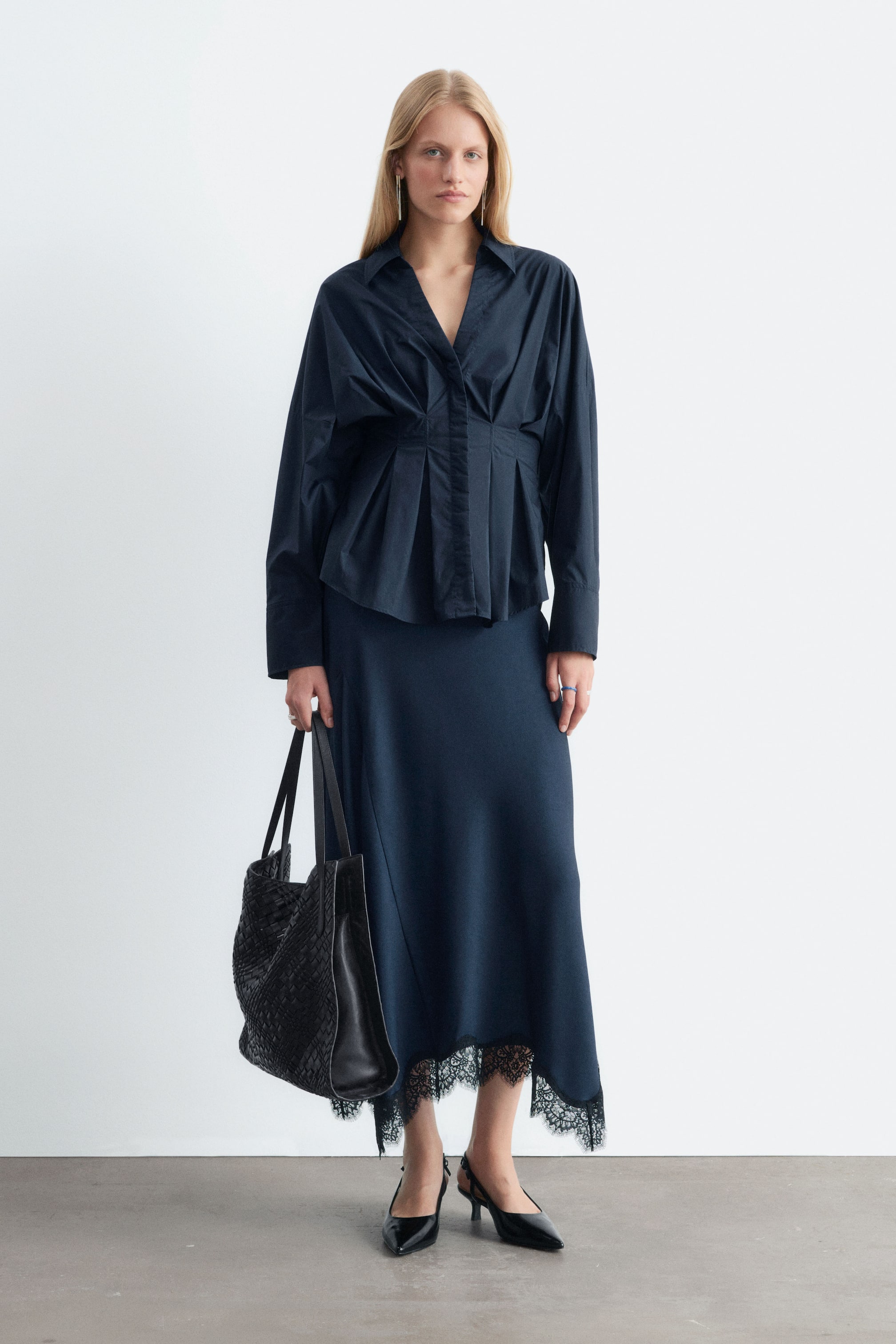 Ver imagen más grande: La modelo lleva una falda acampanada azul oscuro con bajo asimétrico rematado con delicado encaje negro. La prenda se combina con una blusa plisada de manga larga en azul oscuro a juego, unos zapatos de tacón slingback negros y un bolso tote negro de tejido trenzado.