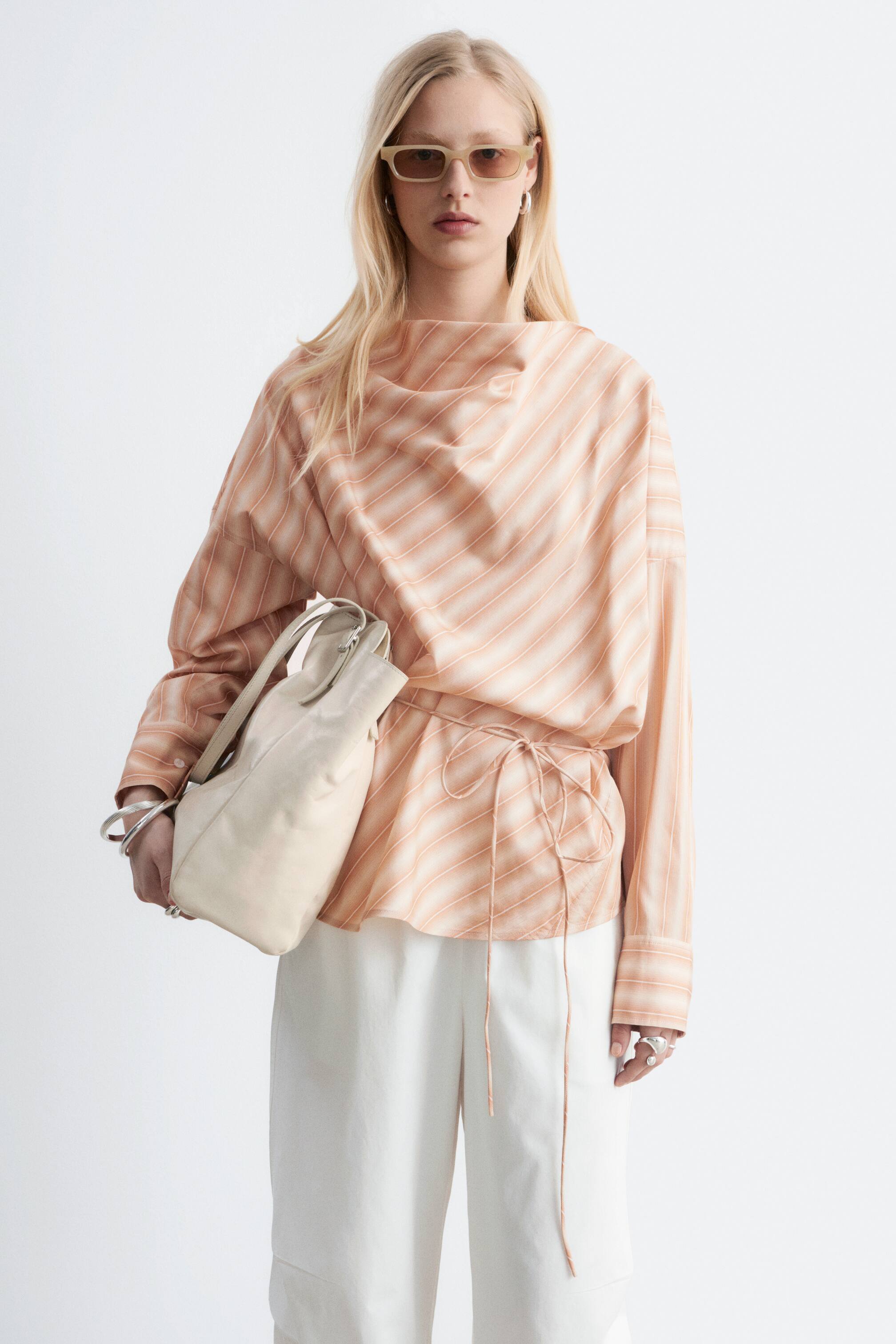 Ingrandisci l'immagine: Una blusa a righe color pesca chiaro e bianco presenta un ampio scollo drappeggiato e maniche lunghe e rilassate. È stretta in vita da un laccio dello stesso tessuto, creando pieghe e un orlo svasato sotto il laccio.
