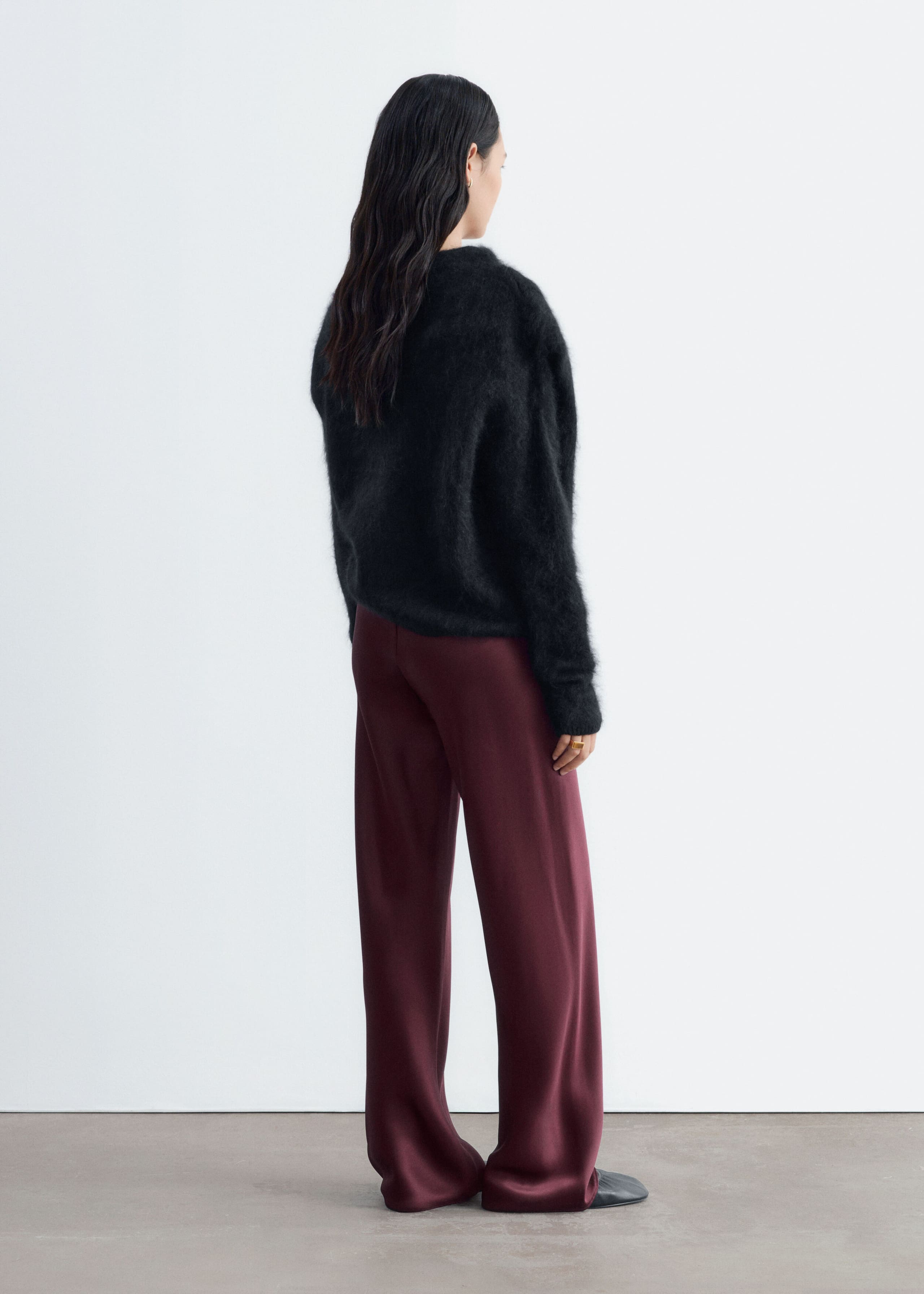 Burgundy satin trousers featuring a relaxed, wide-leg silhouette are worn by the model, who is standing in a three-quarter back view. The fluid, full-length trousers drape to the floor and are styled with a black fuzzy long-sleeved sweater.