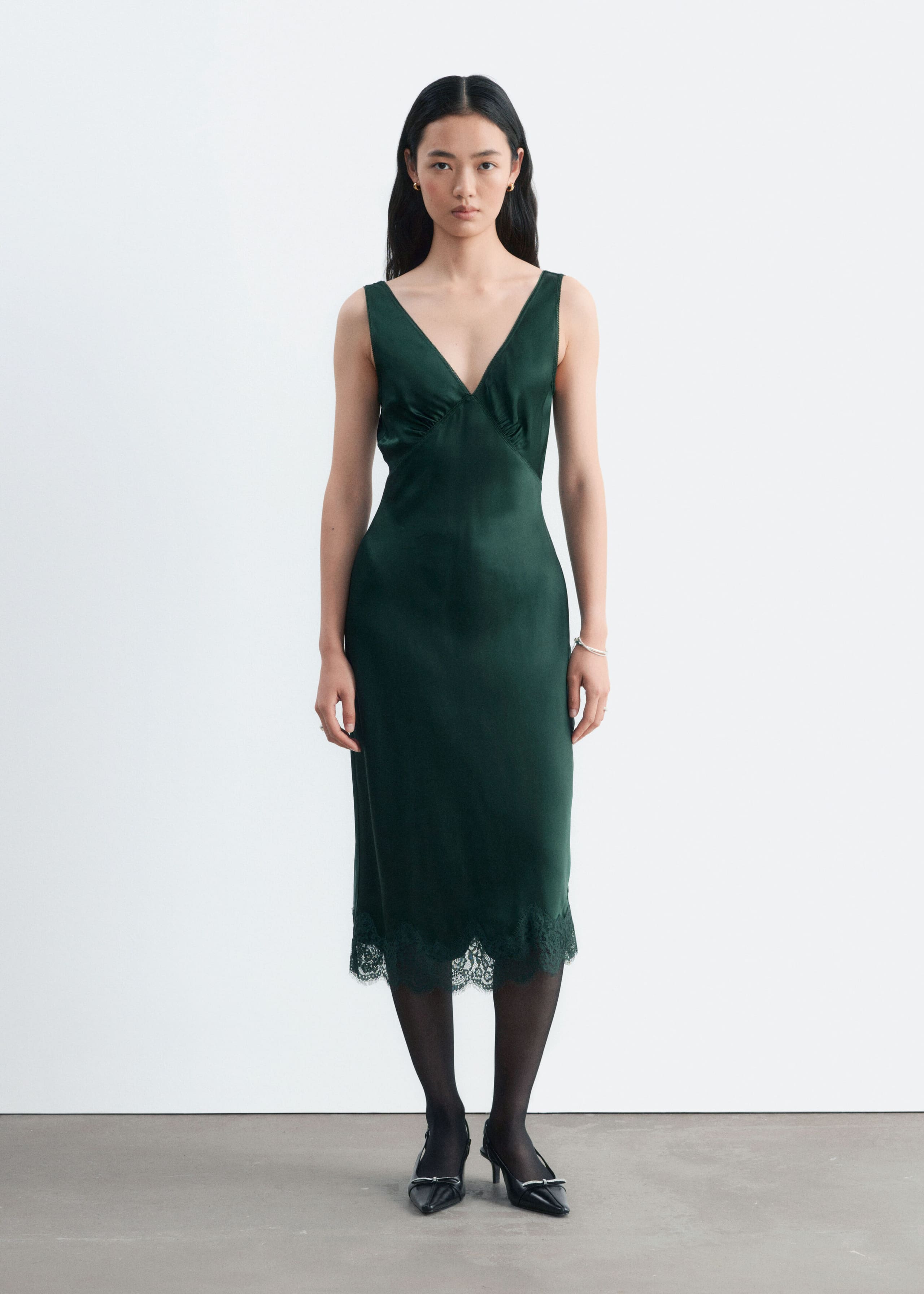 The model is wearing a dark green satin slip dress featuring a V-neckline, gathered bust, and scalloped lace hem. The dress is styled with sheer black tights and black pointed-toe heels.