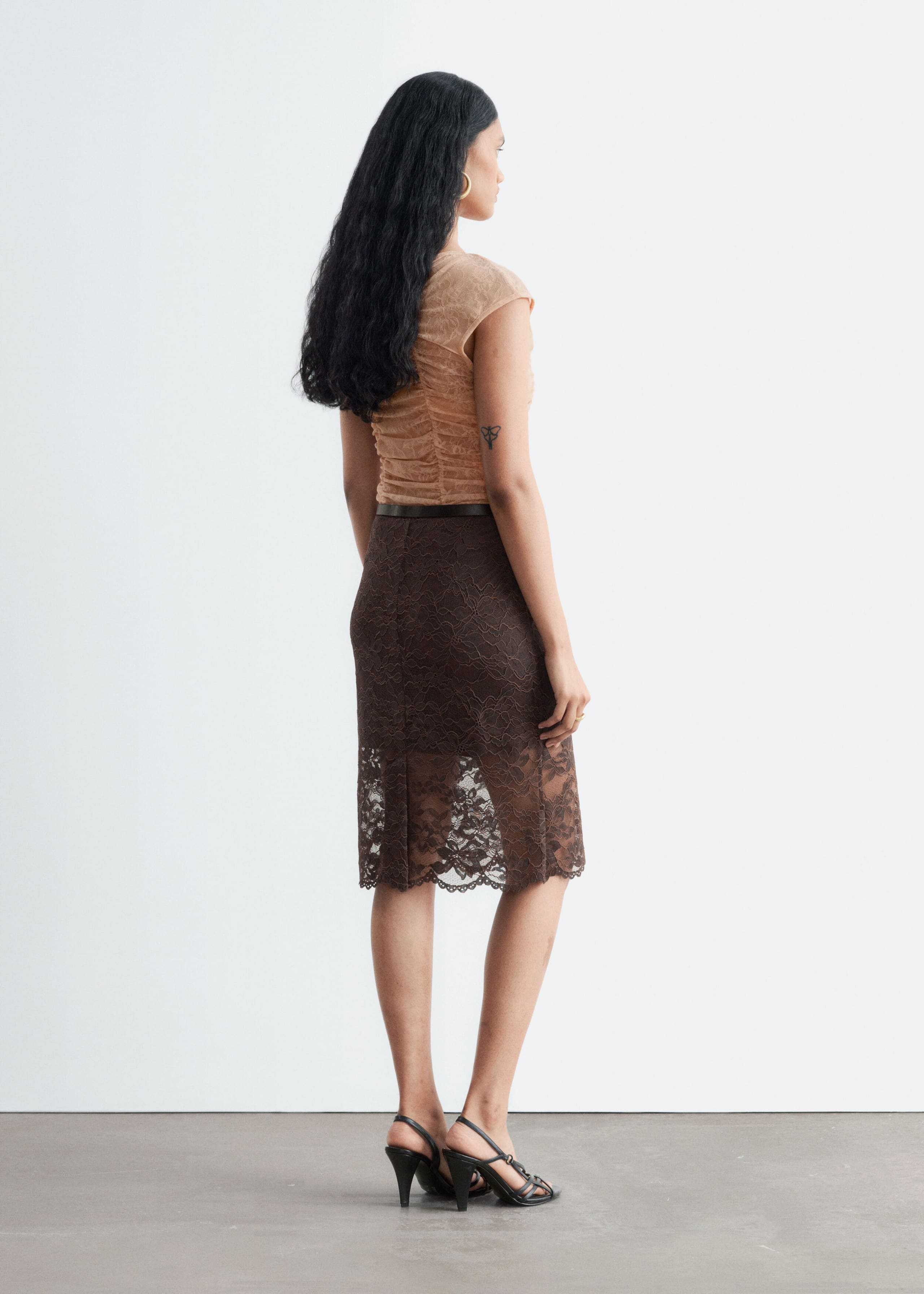 A beige lace top featuring ruching through the body and cap sleeves is being worn by the model, tucked into a dark brown lace skirt. The model is standing in a three-quarter rear view to showcase the fit and texture of the top.