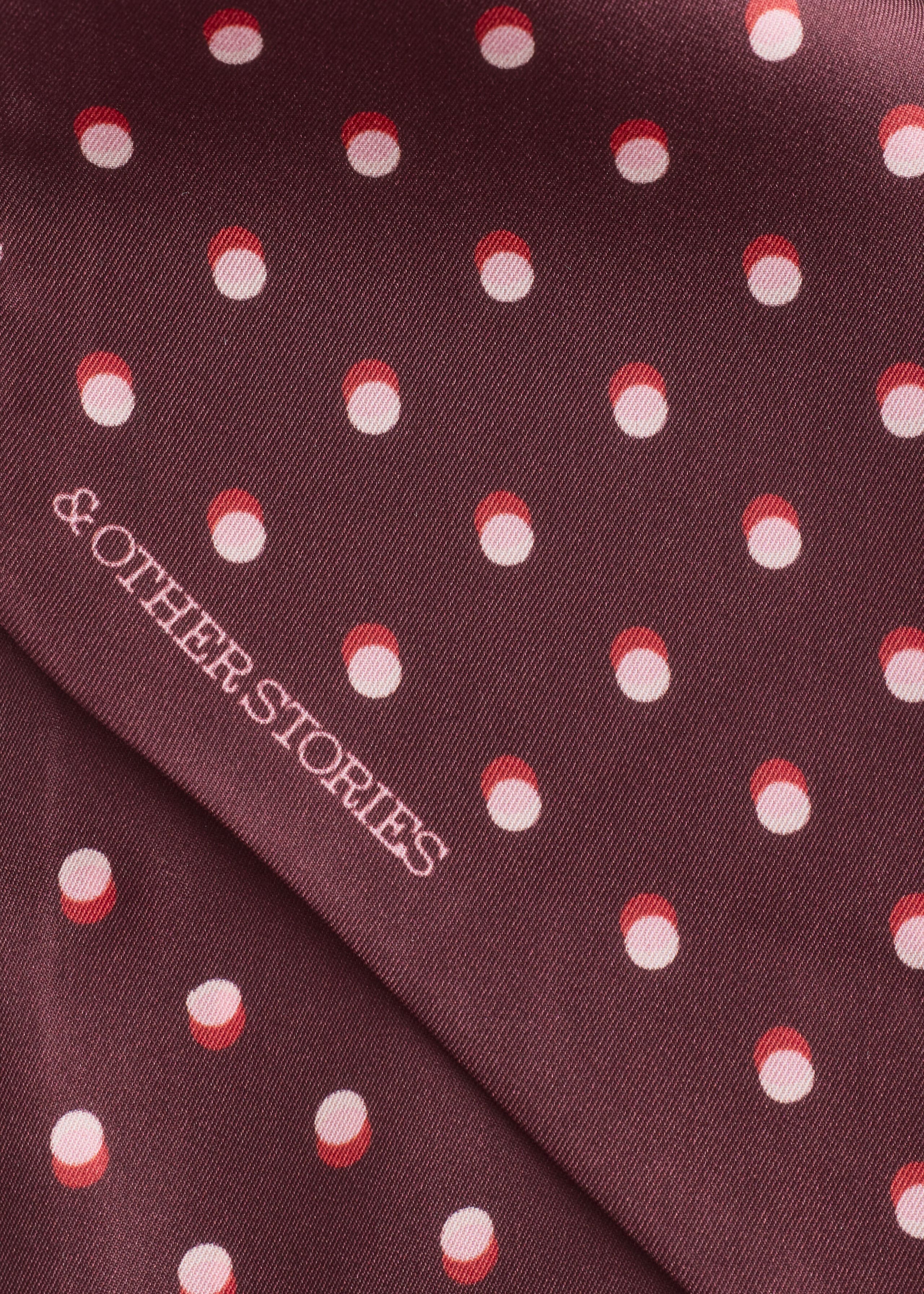 A close-up of a burgundy silk scarf shows an all-over pattern of two-tone polka dots in red and light pink. The brand name “& OTHER STORIES” is printed diagonally across the smooth fabric in a light pink serif font.