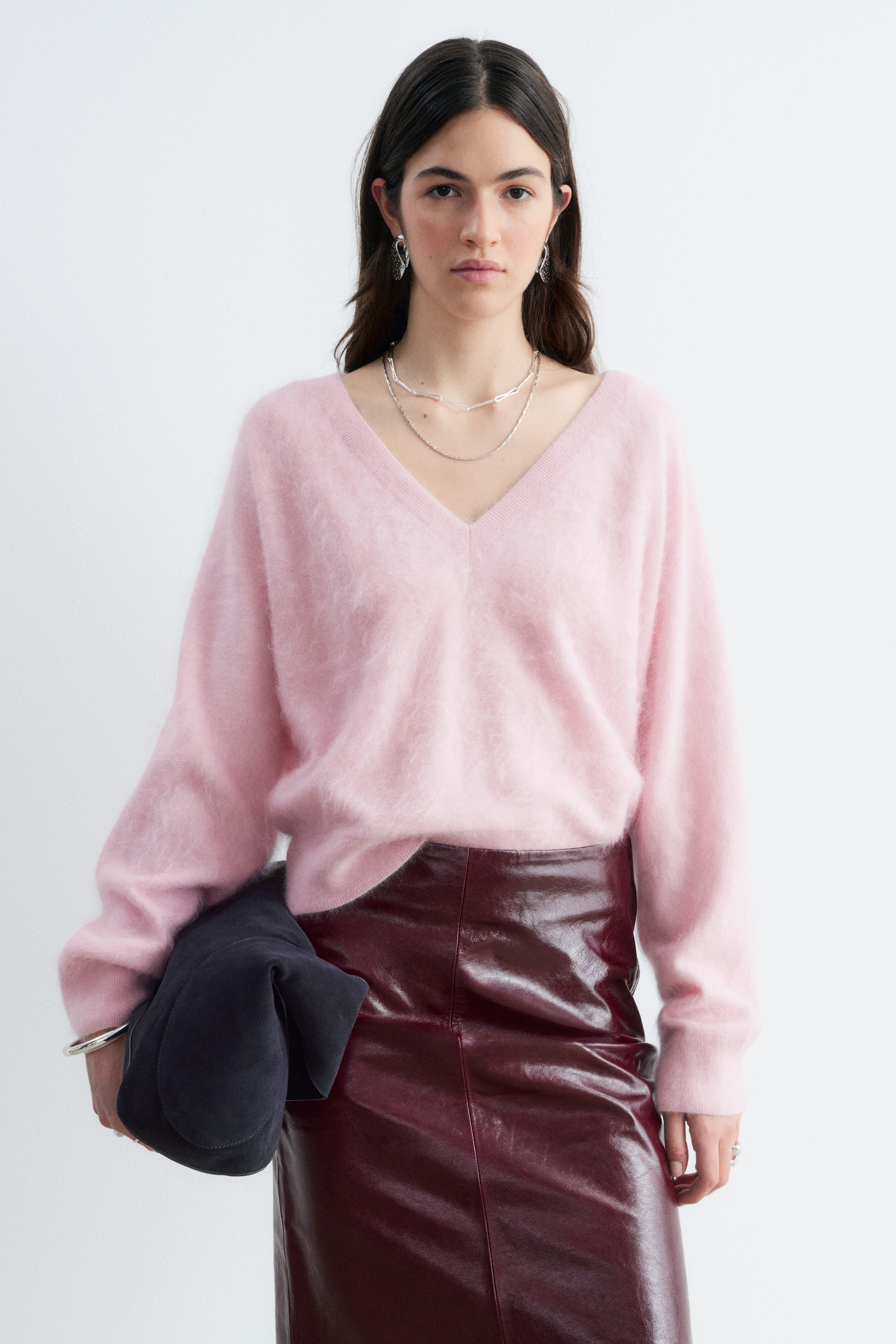 View larger image: A light pink brushed cashmere jumper featuring a fuzzy texture and a V-neckline is being worn by the model, tucked into a glossy, dark red leather skirt. The long-sleeved knitwear is styled with silver chain necklaces, with a dark suede clutch bag held under the model's arm.