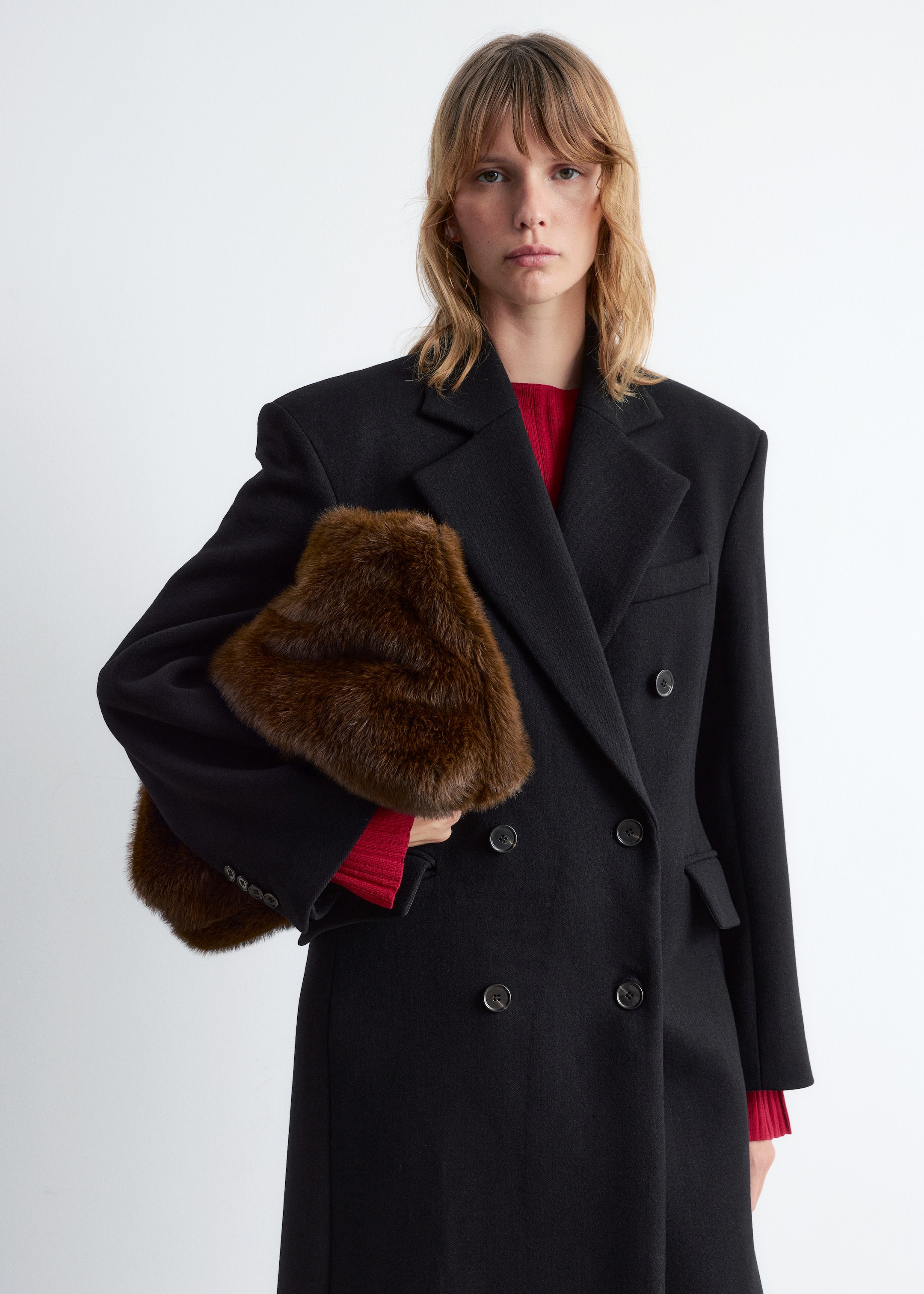 View larger image: A black double-breasted coat is being shown from a slight angle, featuring structured shoulders, notch lapels, and two rows of dark buttons. A red top is visible underneath the coat, and a textured brown fur accessory is being held against the coat's front.