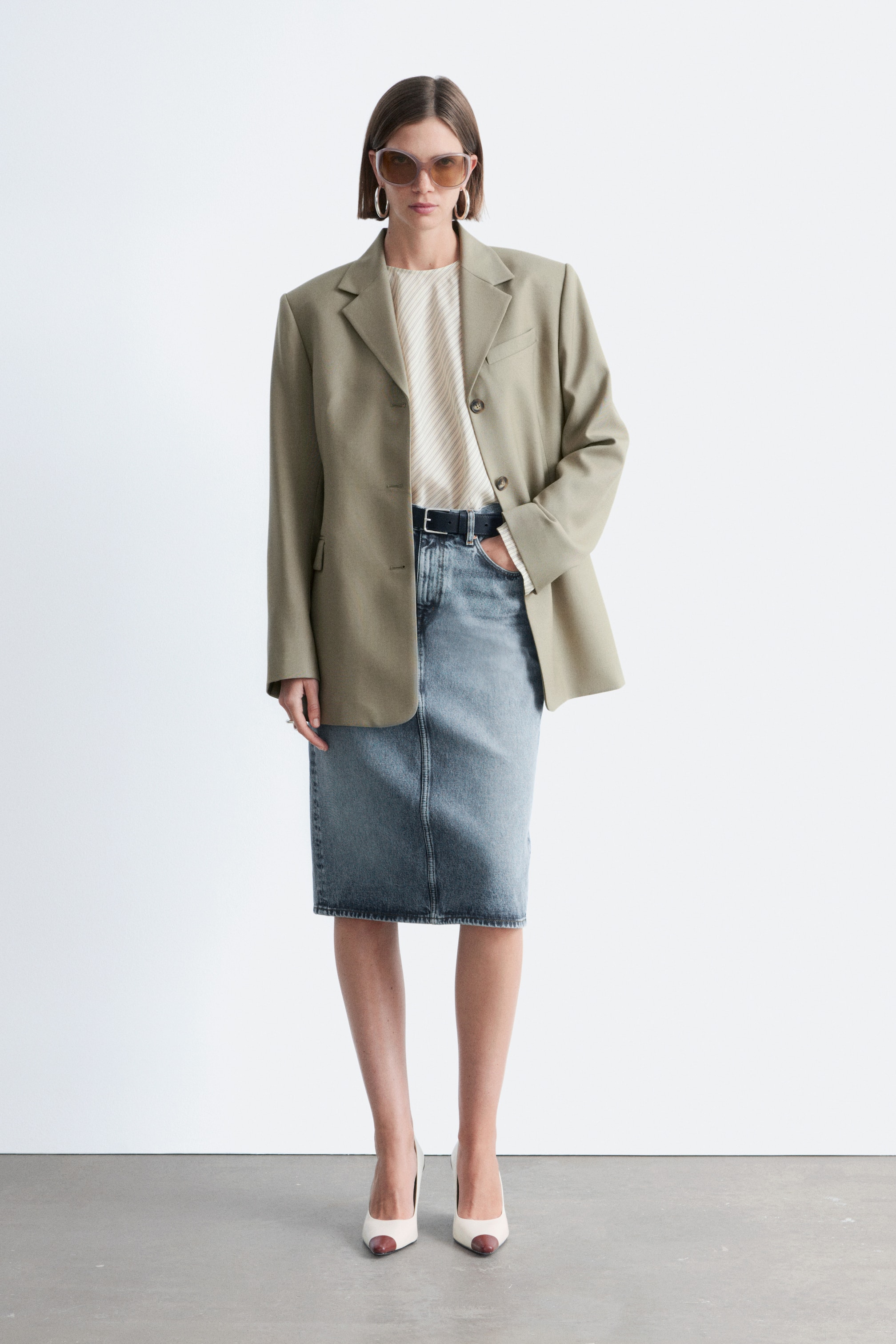 Grotere afbeelding bekijken: Een oversized licht olijfkleurige blazer met inkeping revers en twee knopen wordt open gedragen over een crèmekleurige top met diagonale strepen. Het is gecombineerd met een lichtgewassen denim midi-rok, gestyled met een zwarte riem en tweekleurige pumps.