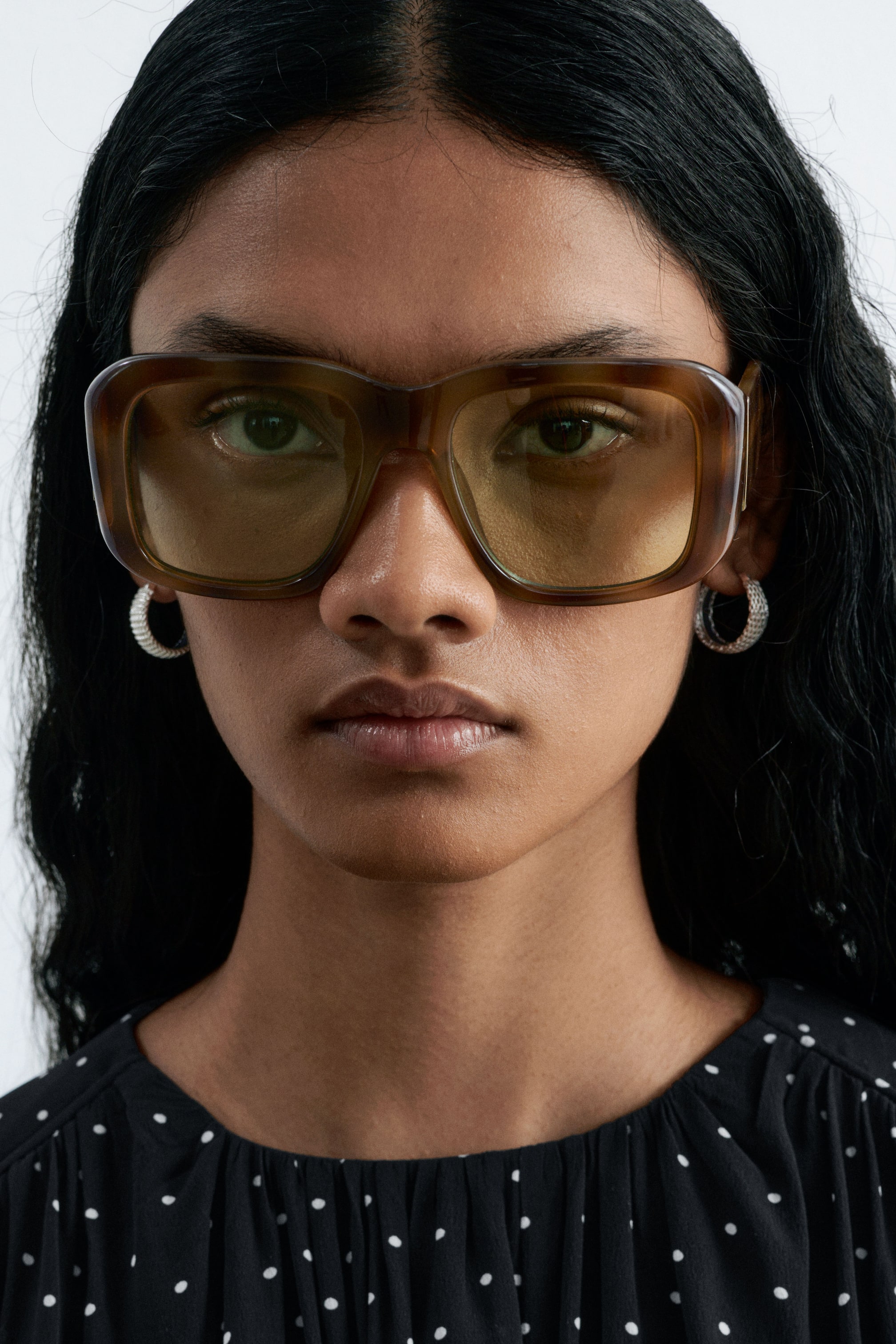 View larger image: Chunky, square-framed sunglasses made of brown tortoiseshell acetate with tinted yellow lenses are worn by the model. The sunglasses are styled with textured silver hoop earrings and a black polka-dot top.