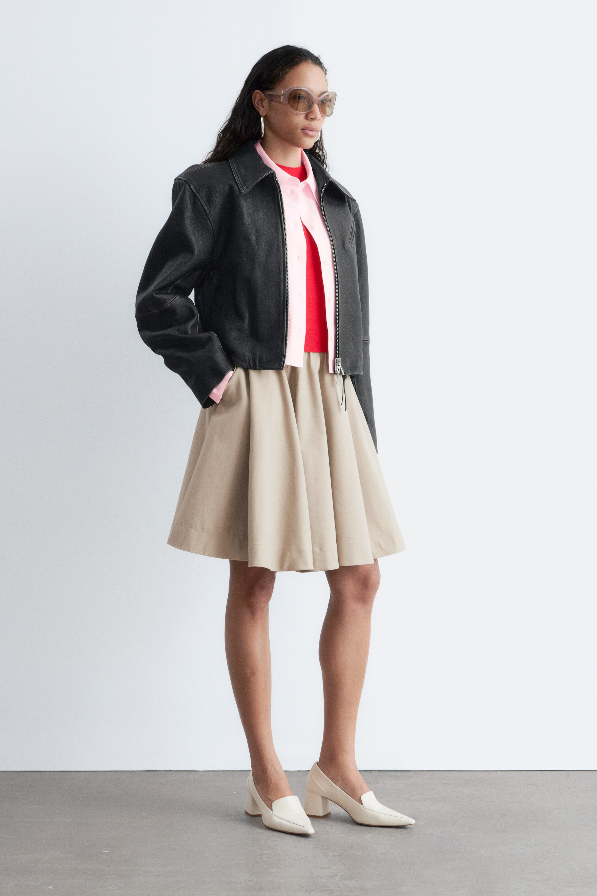 Nagyobb kép megtekintése: A modell egy fekete, rövidített bőrkabátot visel, amely matt, texturált felülettel, kiemelkedő válltömésekkel és dobozszerű sziluettel rendelkezik, rózsaszín galléros ing és piros felső fölött. A modell egyik kezét a kabát oldalzsebébe helyezi.