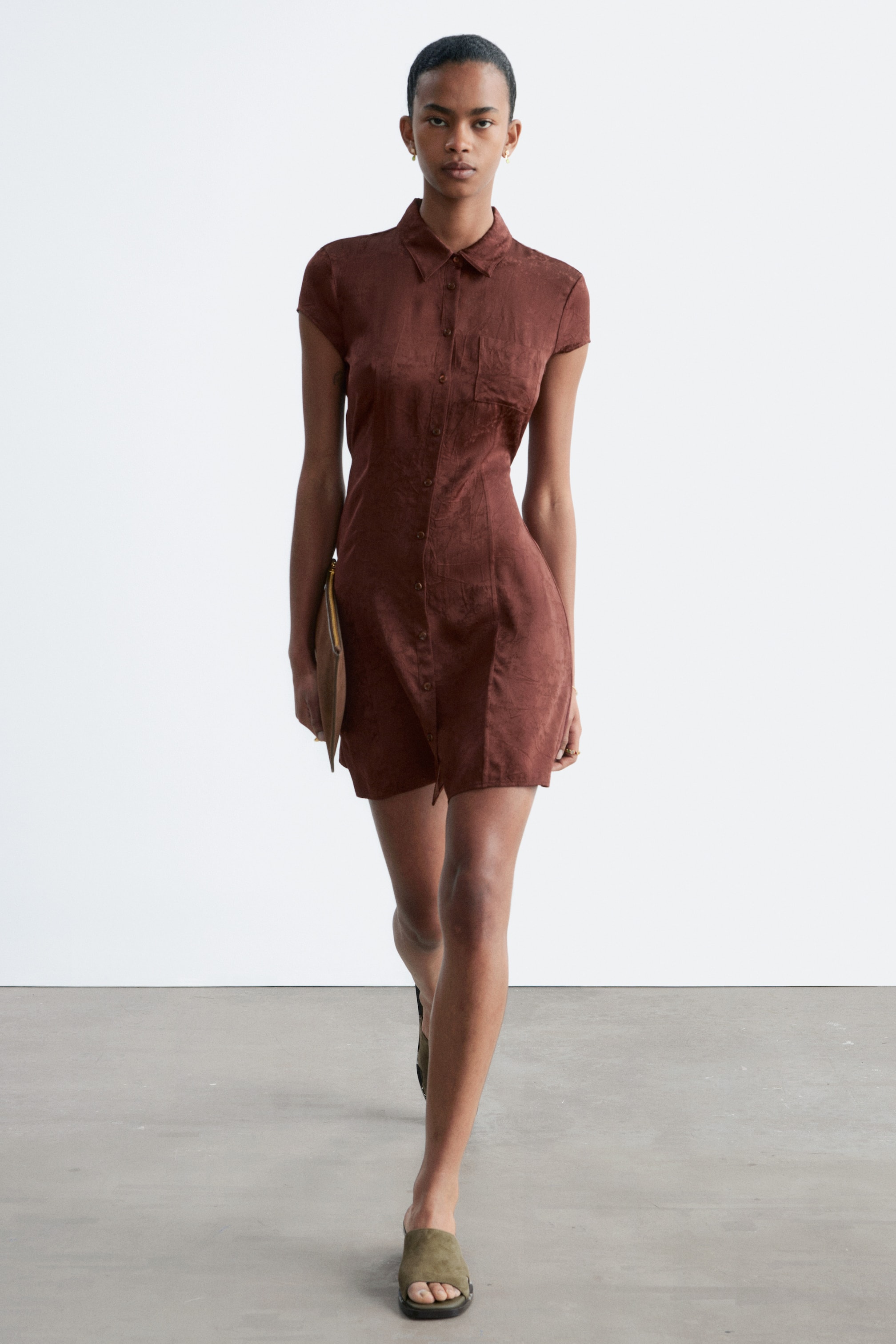 Ver imagem maior: Um vestido-camisa castanho texturizado está virado para a frente, apresentando uma gola pontiaguda, mangas curtas, uma carcela completa com botões à frente e um único bolso aplicado no peito esquerdo. Exibe uma silhueta ajustada que termina acima do joelho.