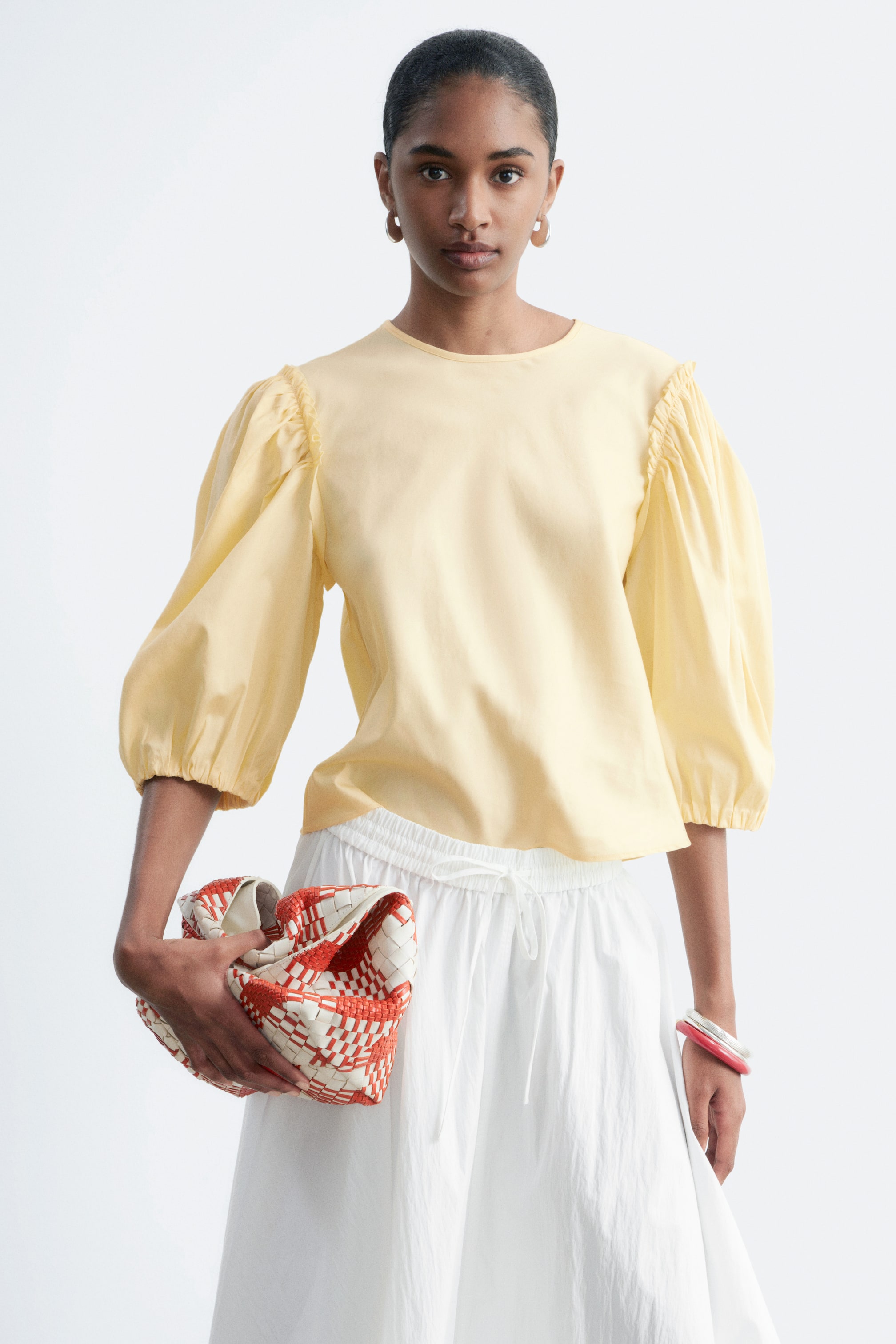 Ver imagen más grande: Una blusa de color amarillo claro, con un escote redondo, hombros fruncidos y mangas abullonadas voluminosas con puños elásticos, es llevada por la modelo. La modelo combina la blusa con una falda blanca con cordón y sostiene un bolso de mano tejido en rojo y blanco.