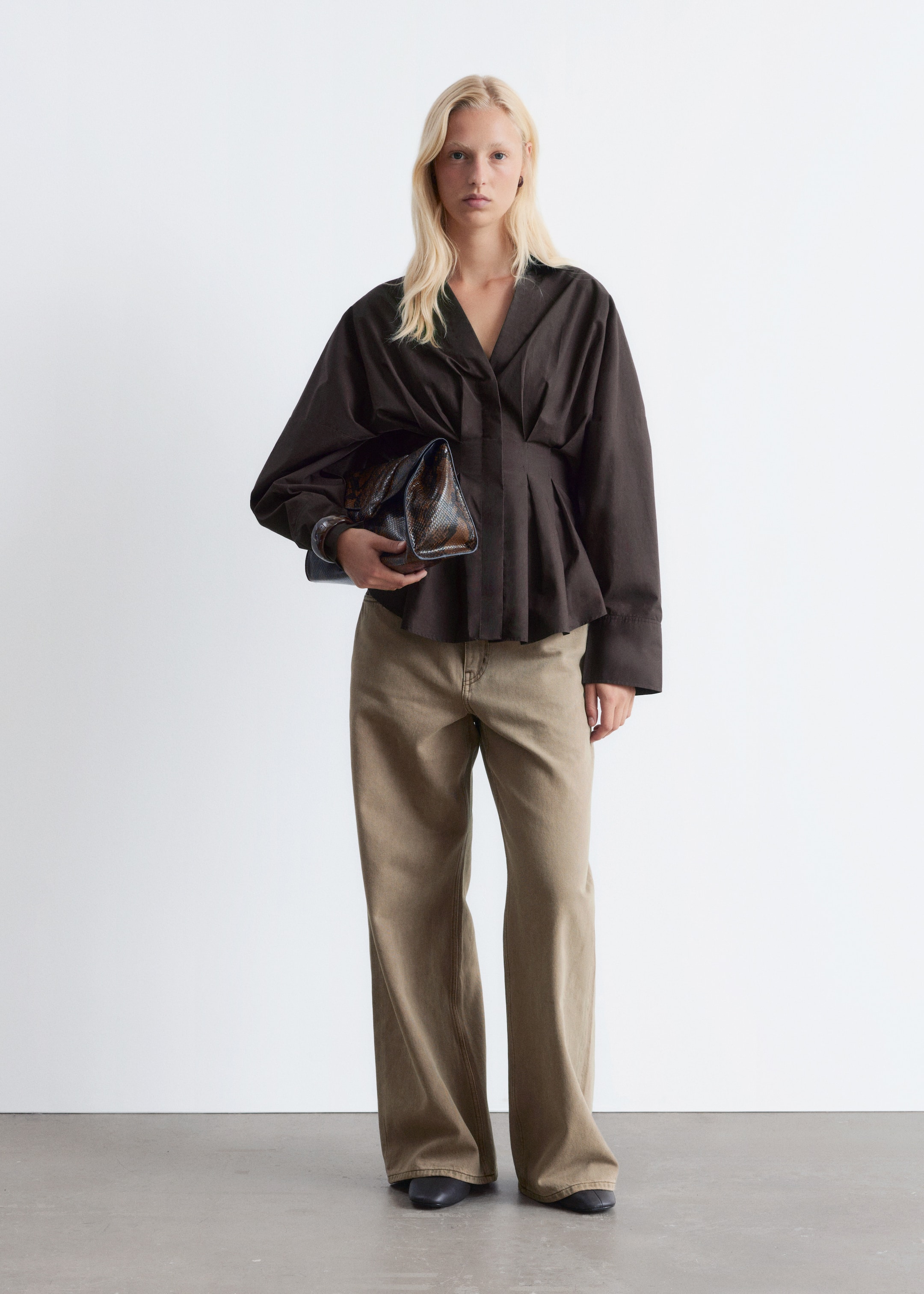 Agrandir l'image: Un haut marron foncé à manches longues, avec un décolleté en V, des manches volumineuses et une taille cintrée et plissée qui s'évase en un ourlet péplum, est présenté. Il est stylisé avec un pantalon large de couleur marron clair et des chaussures plates foncées, tandis qu'une pochette à motif brillant et foncé est tenue dans la main gauche, et un bracelet épais de couleur claire est visible sur le poignet gauche.