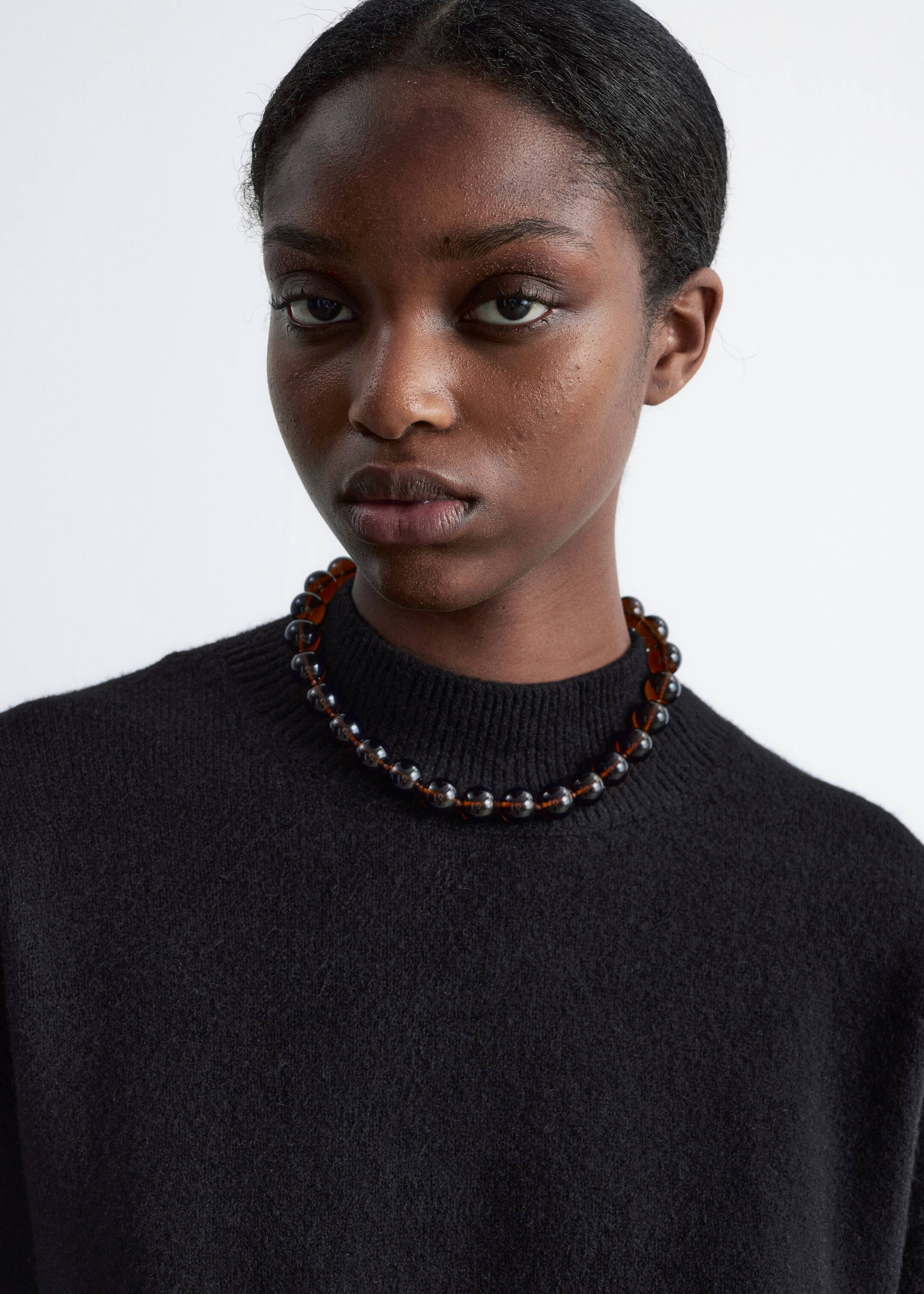 Größeres Bild anzeigen: Eine Halskette im Choker-Stil mit großen, runden, durchscheinenden braunen und dunkelgrauen Perlen, die auf einer dünnen, sichtbaren Schnur aufgereiht sind. Die Halskette liegt knapp über dem Schlüsselbein und wird über einem schwarzen Rollkragenpullover getragen.