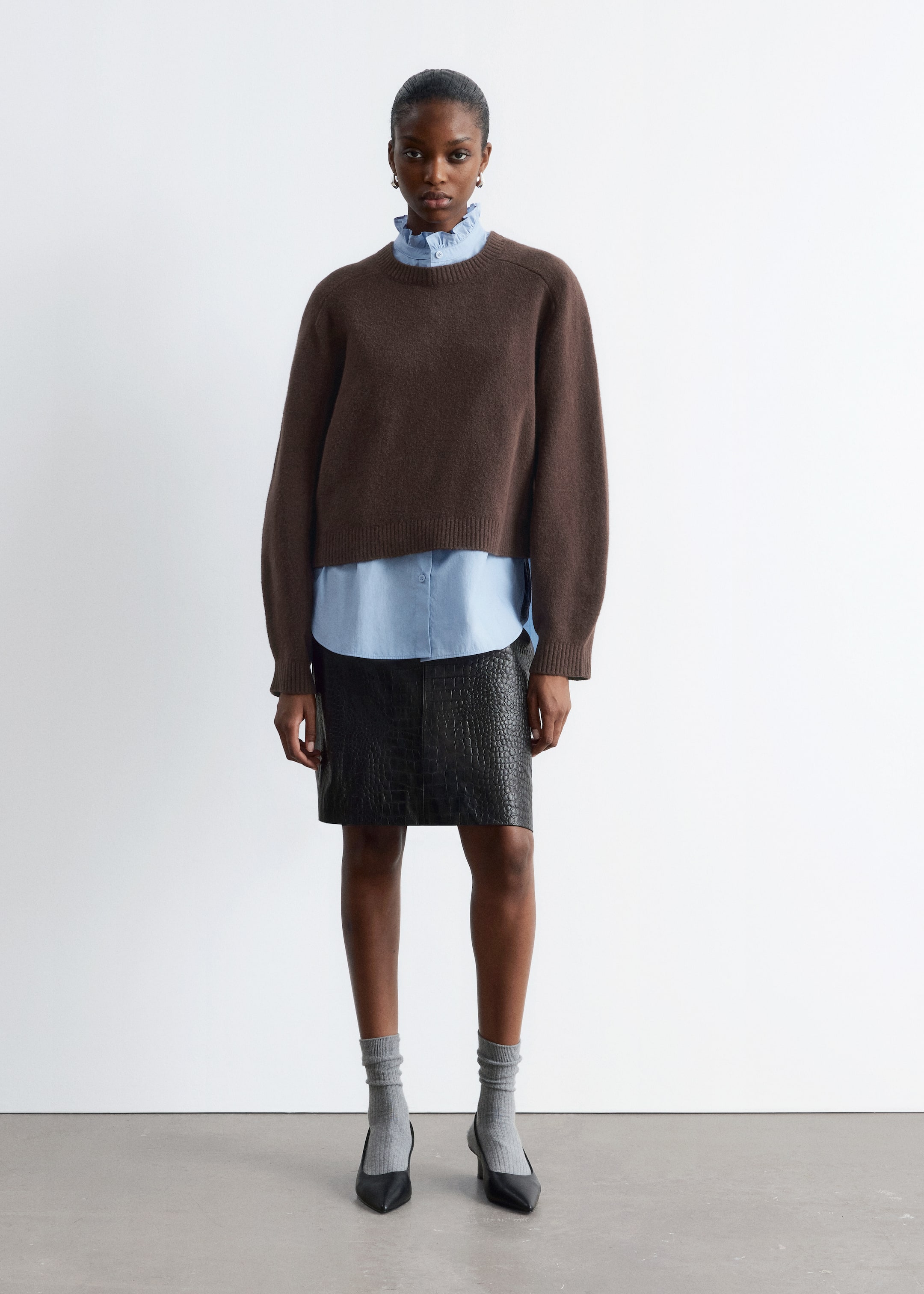 Grösseres Bild ansehen: Ein brauner Pullover mit Rundhalsausschnitt wird getragen, nach vorne gerichtet, mit einer entspannten Passform und langen Raglanärmeln. Er präsentiert eine verkürzte Länge mit gerippten Bündchen und einem gerippten Saum und ist über einem hellblauen Hemd mit Kragen geschichtet.
