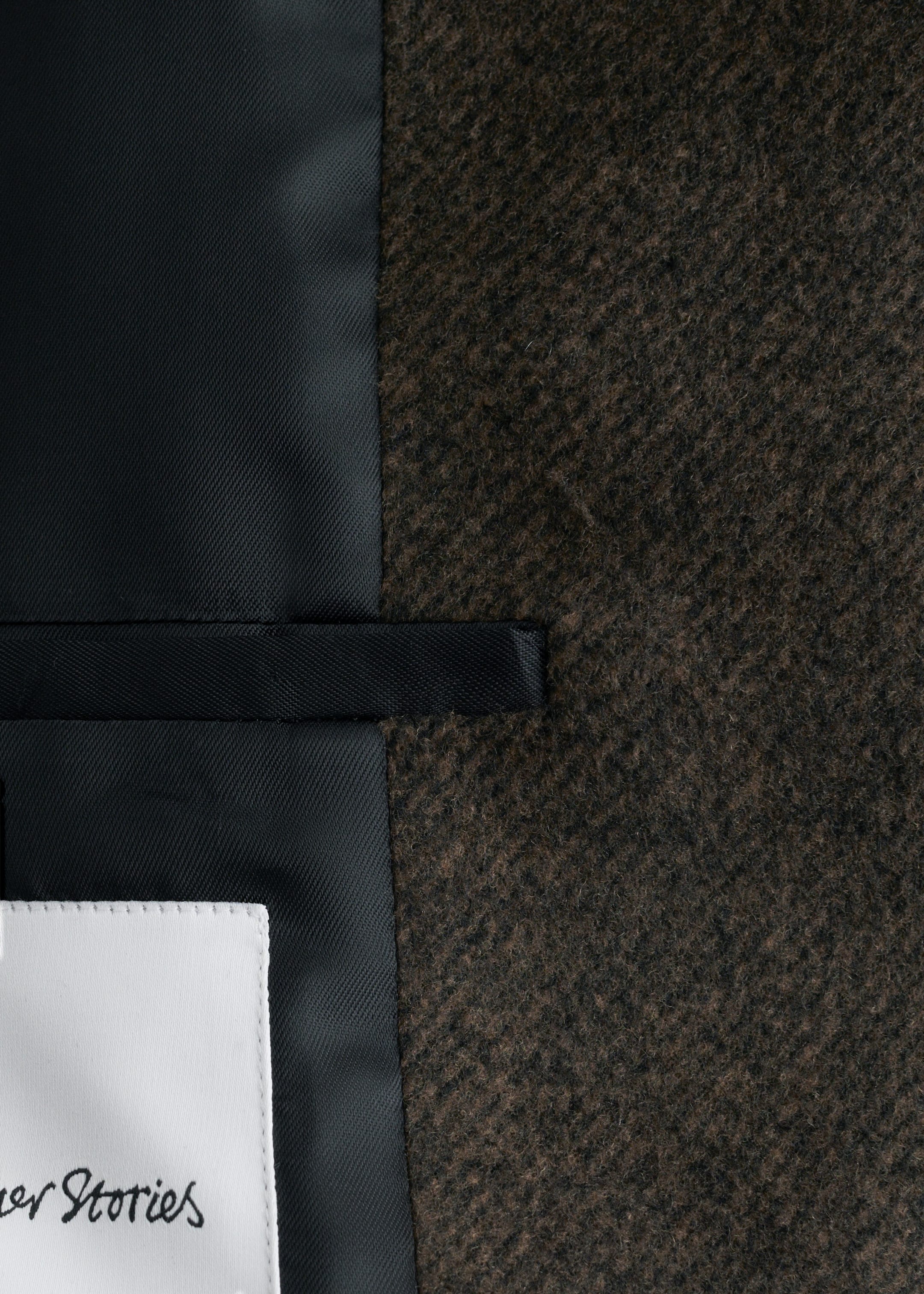 View larger image: The coat's dark brown, textured wool blend exterior is visible on the right, contrasting with the smooth black lining on the left. An internal black strap is connecting the exterior and lining, with a white brand tag partially showing on the lining at the bottom left.