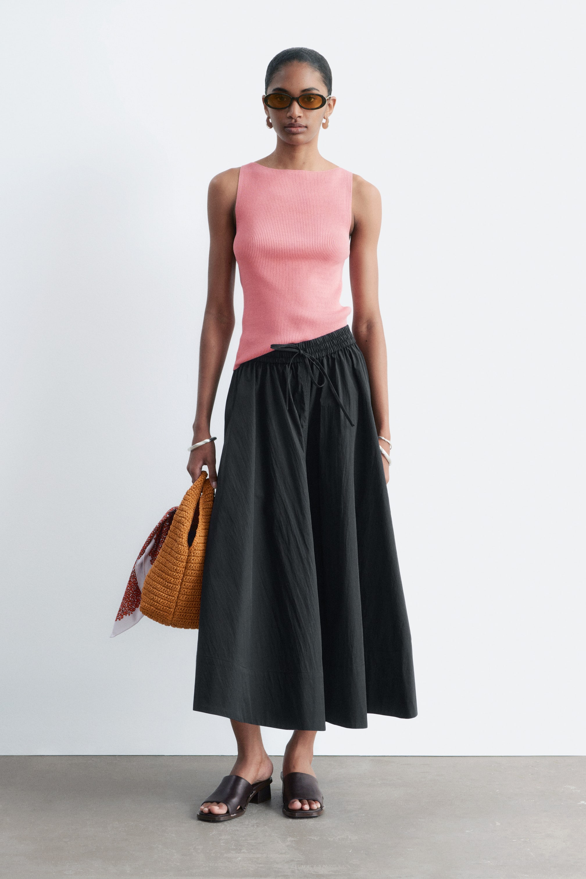 Nagyobb kép megtekintése: Egy fekete maxi szoknya a deréknál van összegyűjtve, látható zsinóros megkötővel, és bőségesen áramlik a bokákig. A modellen a szoknya látható, amelyet egy rózsaszín bordázott tank top és barna papucsokkal stílusosan párosítottak.