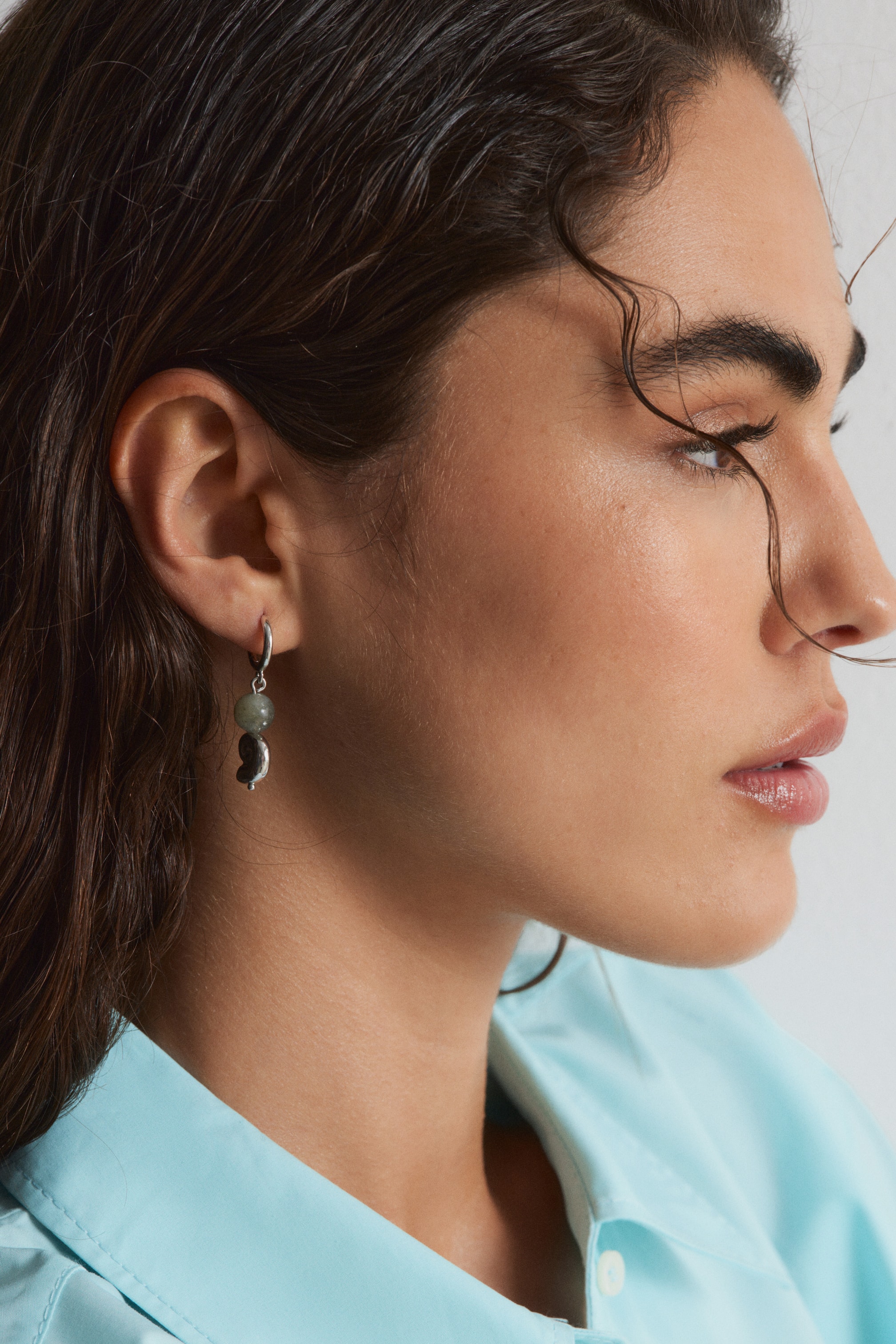 View larger image: A drop earring consisting of a silver-toned hoop, from which a rounded grey-green bead and an irregularly shaped, silver-toned pendant hang, displayed on the wearer's earlobe in a side profile.