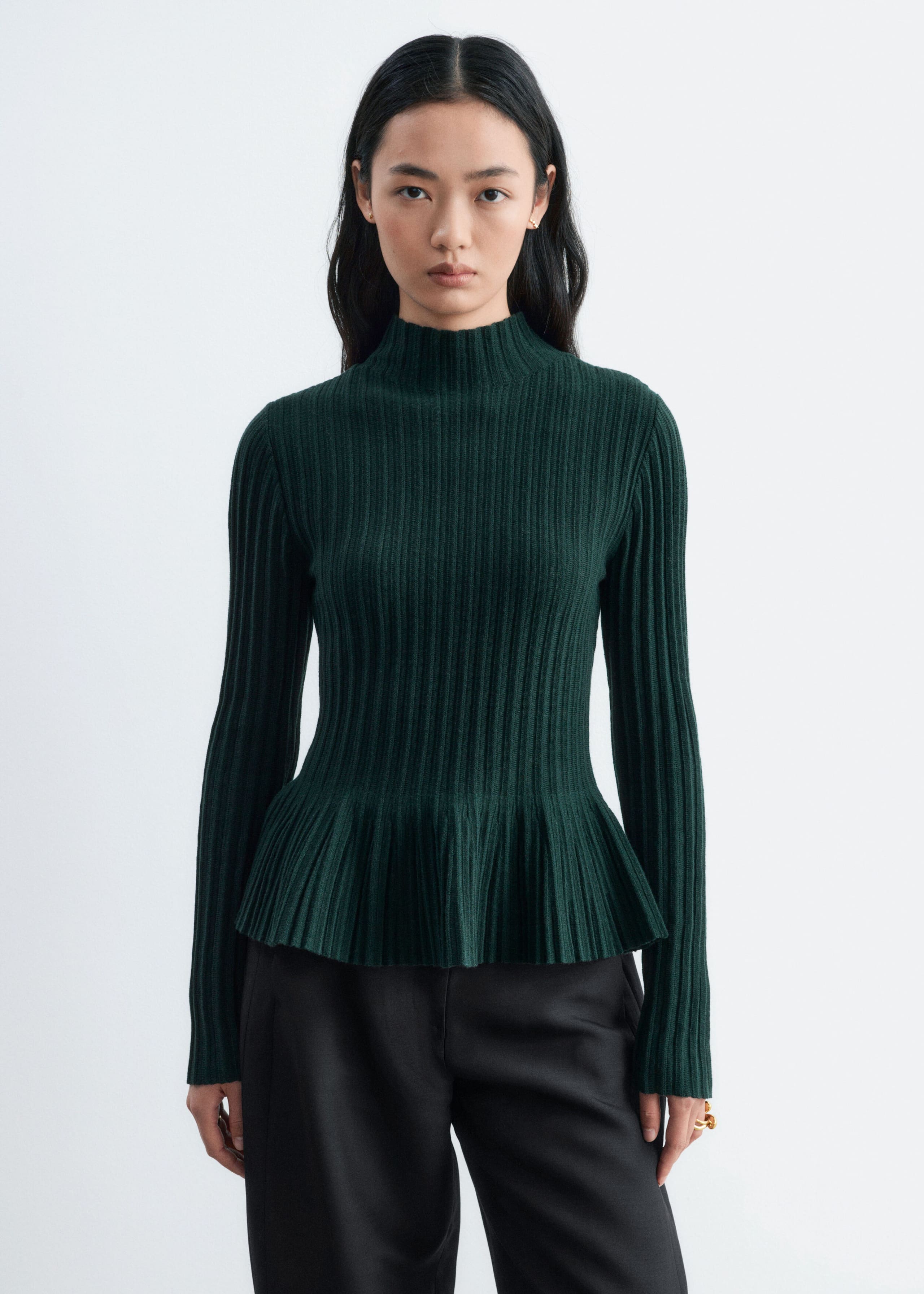 A dark green ribbed-knit top featuring a mock neck, long sleeves and a flared peplum hem is worn by the model. The top is styled with black trousers.