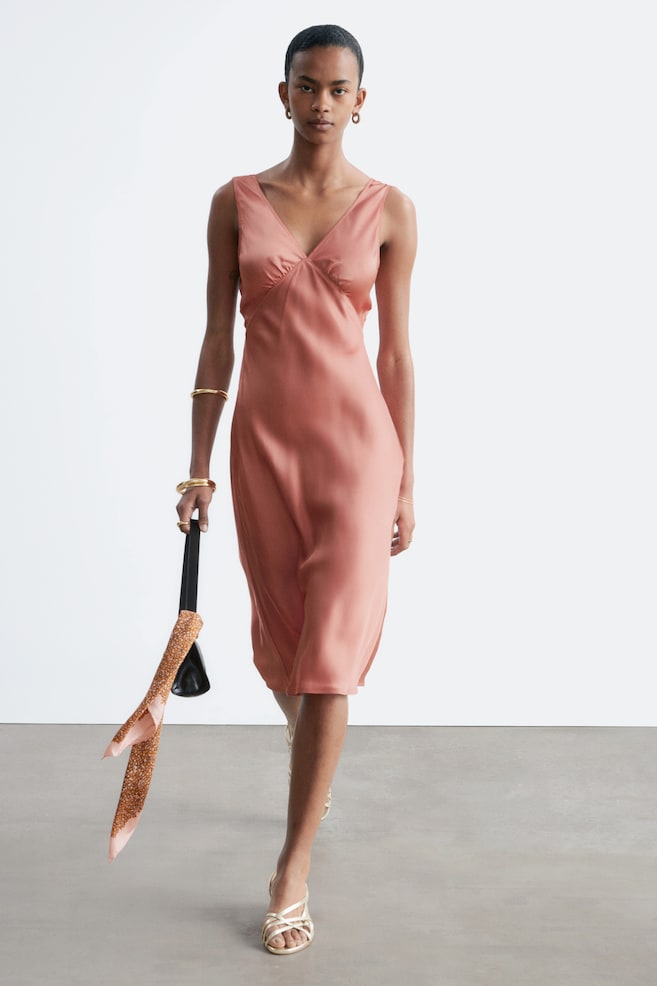 Se lleva un vestido lencero color salmón, ligeramente inclinado hacia la derecha, que destaca un escote en V y detalles fruncidos en el busto. El vestido cae a una longitud midi, con su tejido satinado fluyendo suavemente. - 1