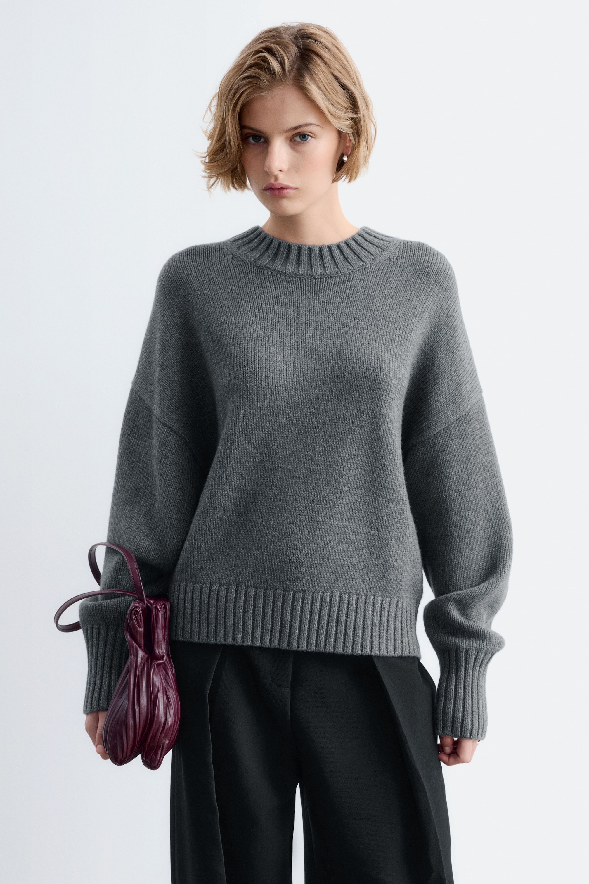 Agrandir l'image: Un pull en maille oversize gris, doté d’un col rond côtelé, d’épaules tombantes et de larges poignets et ourlet côtelés, est porté par le mannequin. Le mannequin associe le pull à un pantalon noir et tient un sac à main en cuir bordeaux foncé.