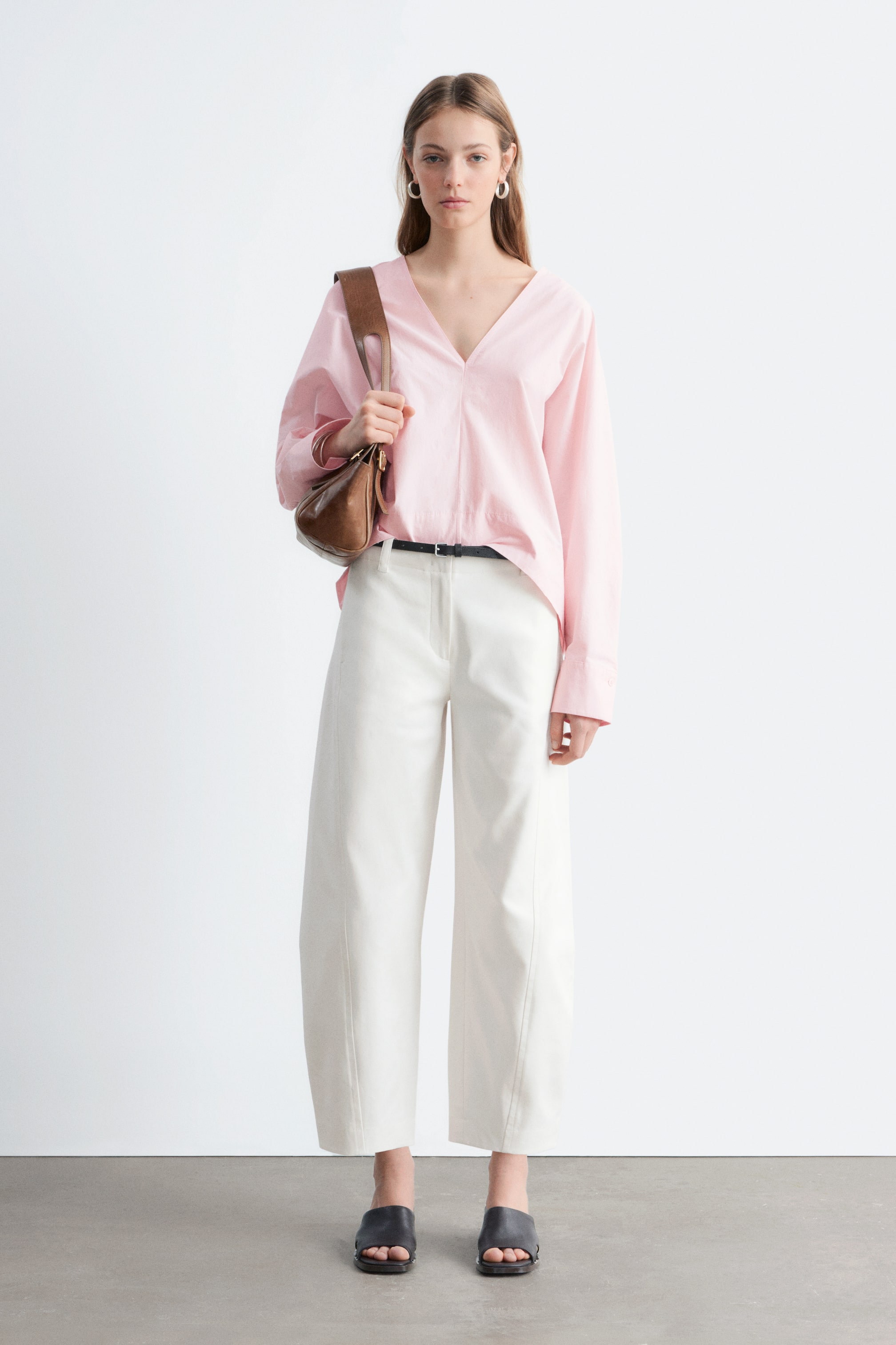 Zobrazit větší obrázek: Světle růžová halenka s výstřihem do V s dlouhými rukávy je kombinována s bílými kalhotami s širokými nohavicemi, které jsou stažené tenkým černým páskem. Černé otevřené pantofle na podpatku doplňují vzhled, a hnědá kabelka ve tvaru půlměsíce je nesena přes rameno.