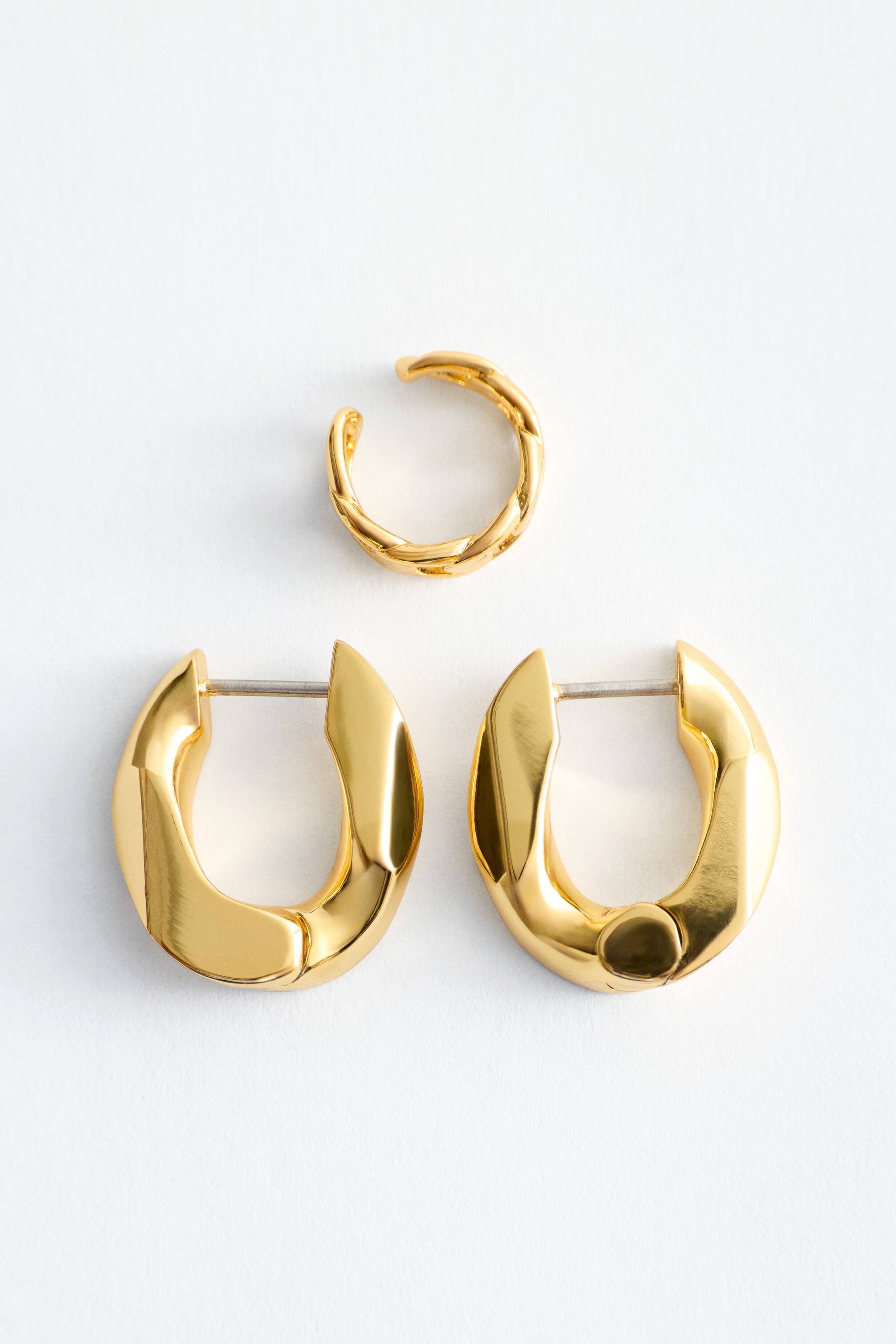 View larger image: Two polished gold-toned sculptural hoop earrings exhibit a bold, curved shape, with one earring showcasing its front latch-back fastening. A smaller chunky chain ear cuff is presented alongside the hoops.