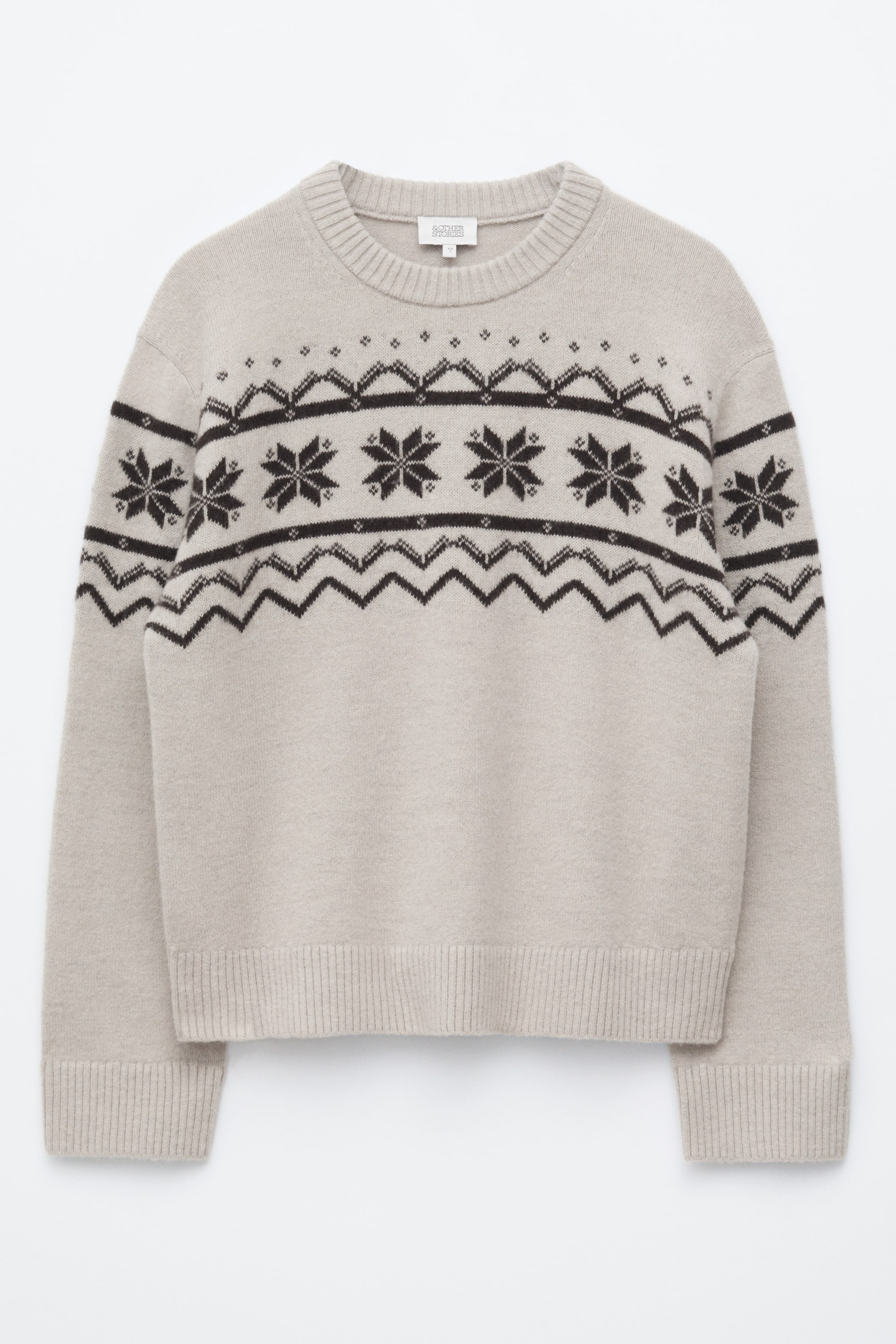 Größeres Bild ansehen: Ein hellbeiger Strickpullover ist von vorne zu sehen und zeigt ein dunkelbraunes Fair-Isle-Muster mit Schneeflocken- und geometrischen Motiven über der Brust und den oberen Ärmeln. Das Kleidungsstück hat eine kastige Silhouette und ist mit einem gerippten Rundhalsausschnitt, gerippten Bündchen und einem gerippten Saum versehen.