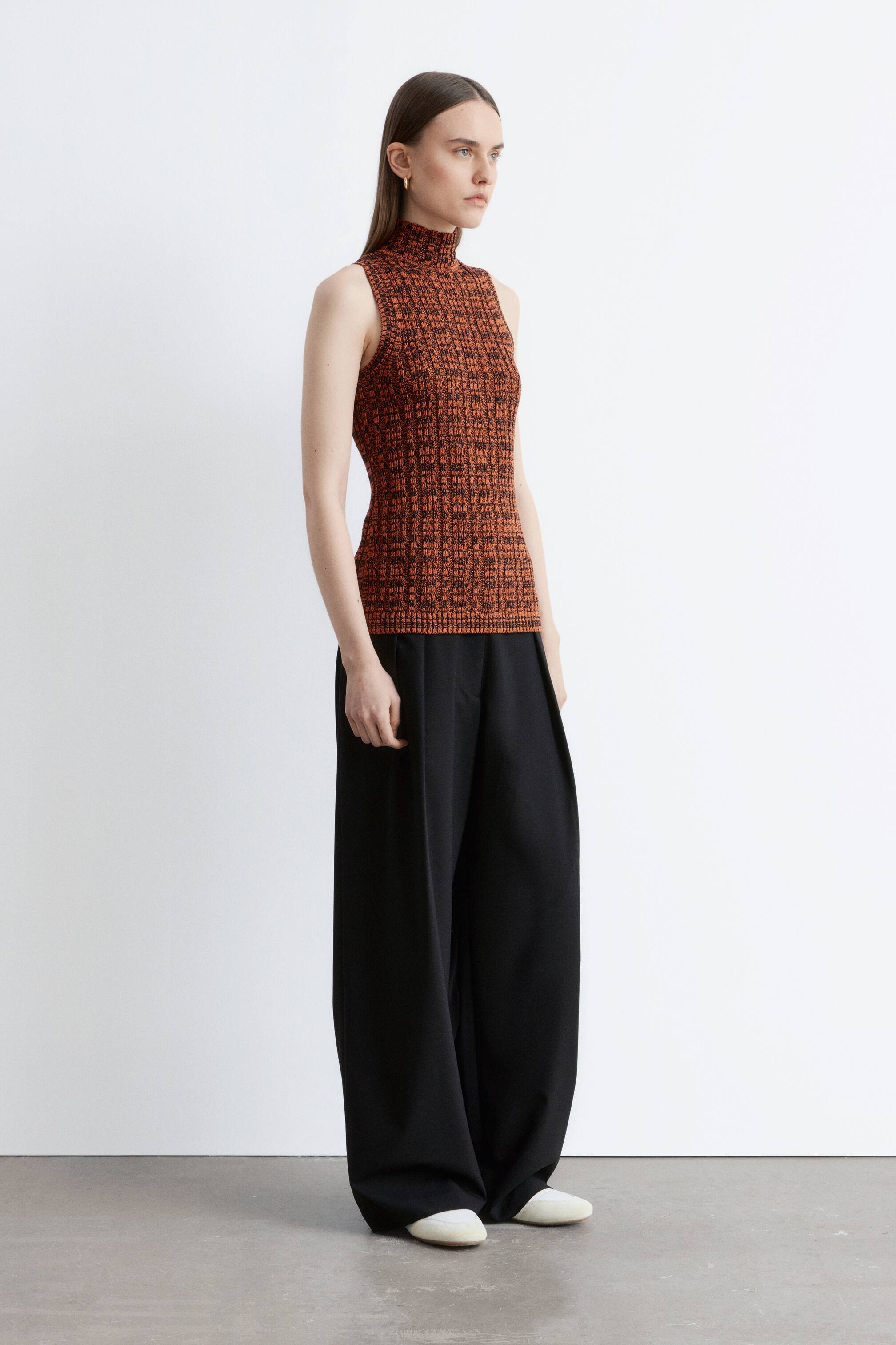 Visa större bild: En ärmlös topp med hög krage i en orange och mörkbrun texturerad stickning med ribbad nederkant visas i sidoprofil. Toppen sitter tätt mot kroppen och är stylad med vida svarta byxor som faller löst.
