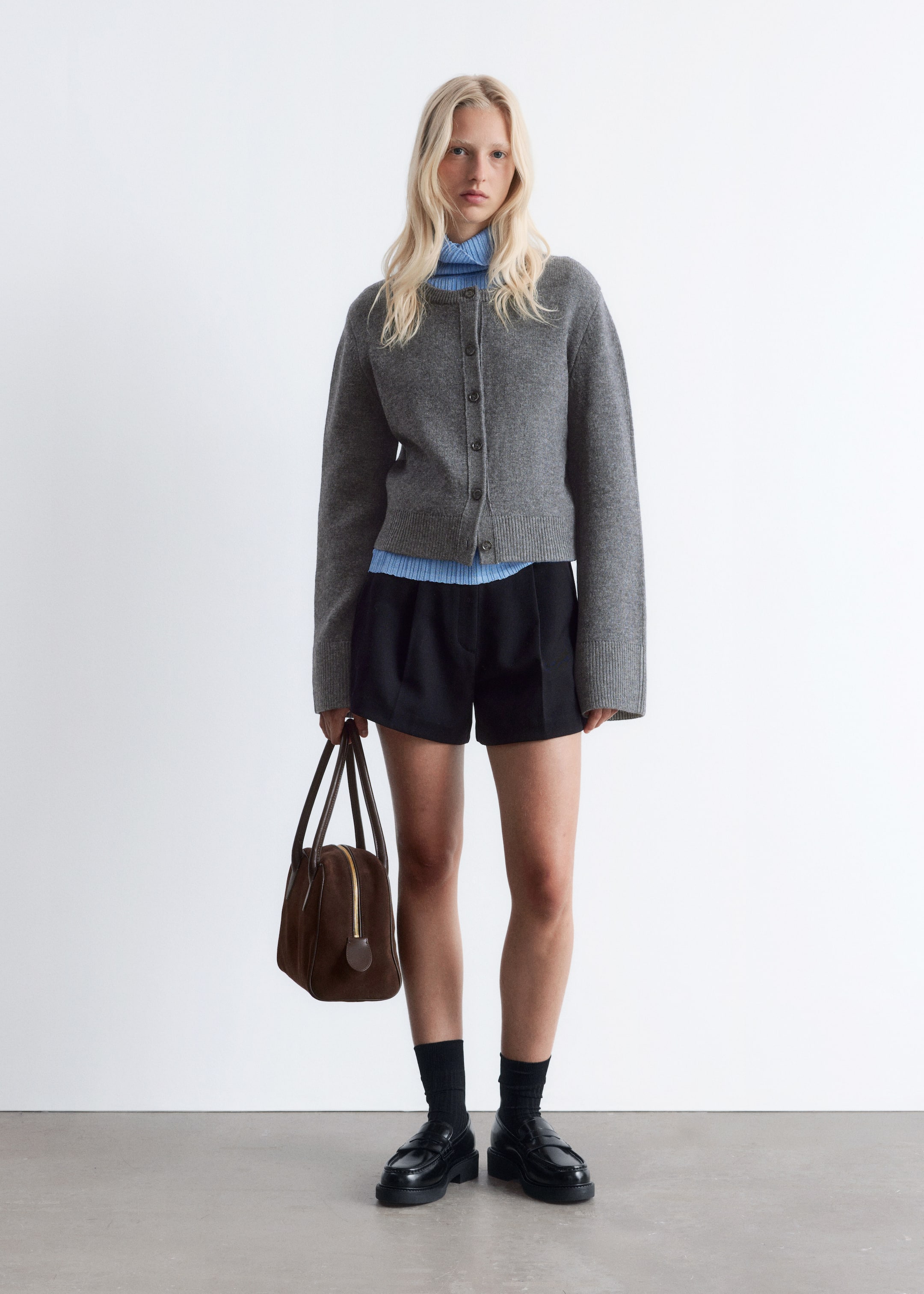 Vis større bilde: En grå cardigan med rund hals og fem knapper bæres over en lyseblå ribbestrikket høyhalset topp, kombinert med svarte plissert shorts, svarte ribbestrikkede sokker og svarte chunky loafers. En brun strukturert veske med topphåndtak holdes i hånden.