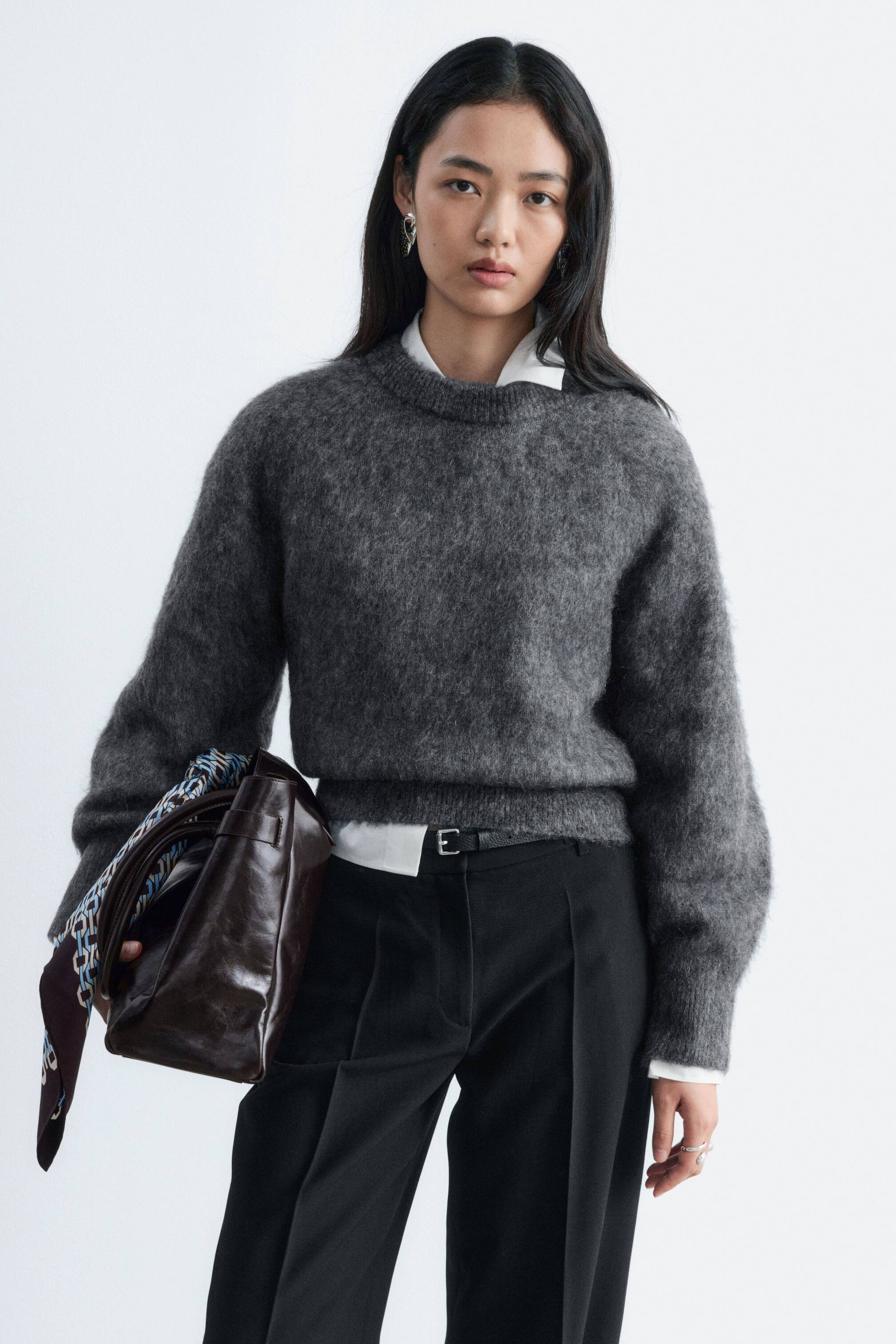 Größeres Bild ansehen: Ein dunkelgrauer, flauschiger Strickpullover wird vom Model getragen und zeichnet sich durch einen runden Rippkragen, Raglan-Ballonärmel und einen breiten gerippten Saum aus. Der Pullover wird über einem weißen Hemd mit Kragen getragen und mit einer schwarzen Hose gestylt, während das Model eine dunkelbraune Ledertasche trägt.