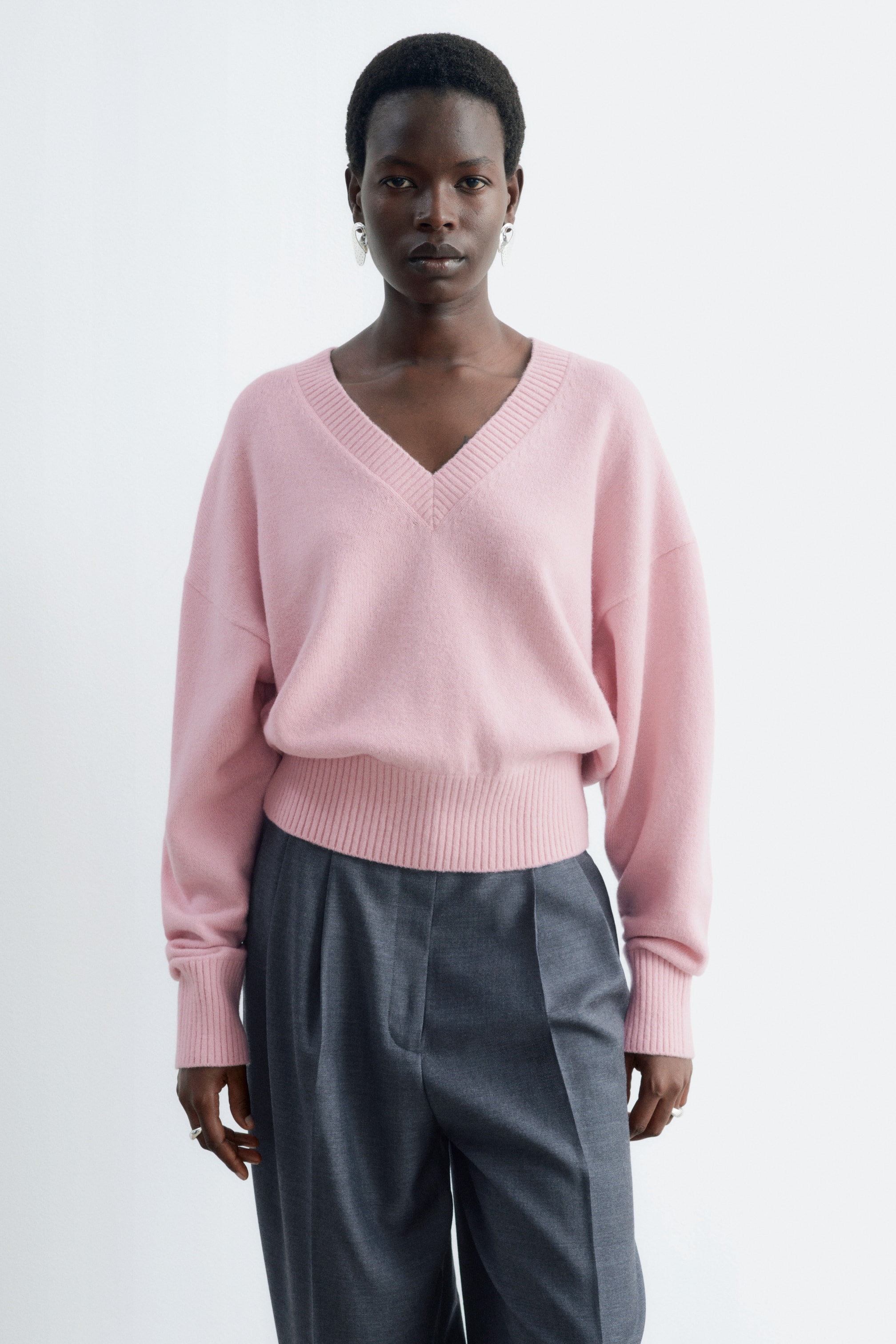 Ver imagen más grande: La modelo lleva puesto un jersey rosa de cuello en V. Presenta hombros caídos, un pronunciado cuello en V y amplios acabados acanalados en el bajo y los puños, con el bajo cayendo por encima de un pantalón gris.