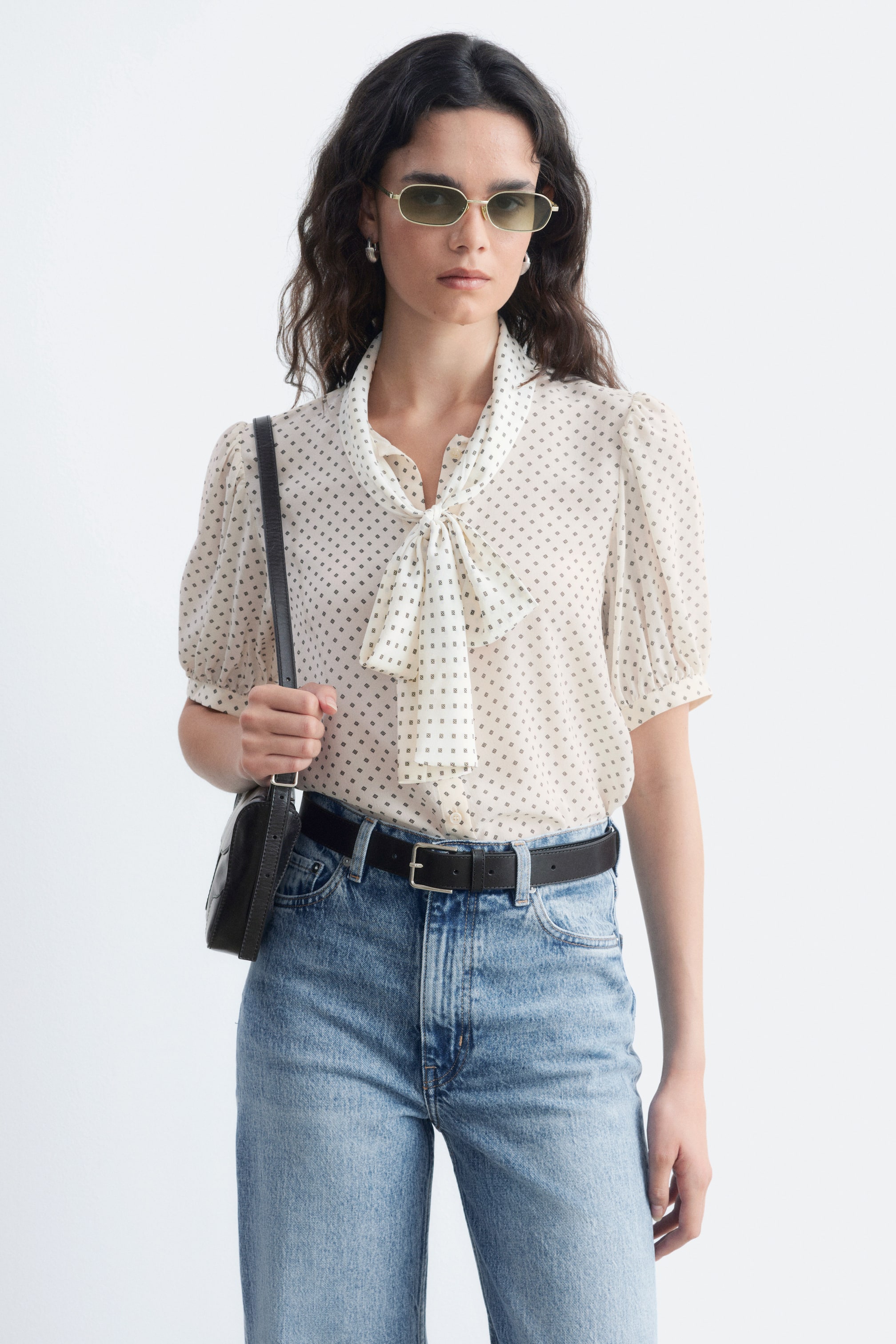 Größeres Bild ansehen: Eine cremefarbene Seidenbluse mit dunklem Mikromuster wird von dem Model getragen, in eine hoch taillierte blaue Jeans mit schwarzem Gürtel gesteckt. Die Bluse hat kurze, geraffte Puffärmel und einen vorne gebundenen Bindekragen.