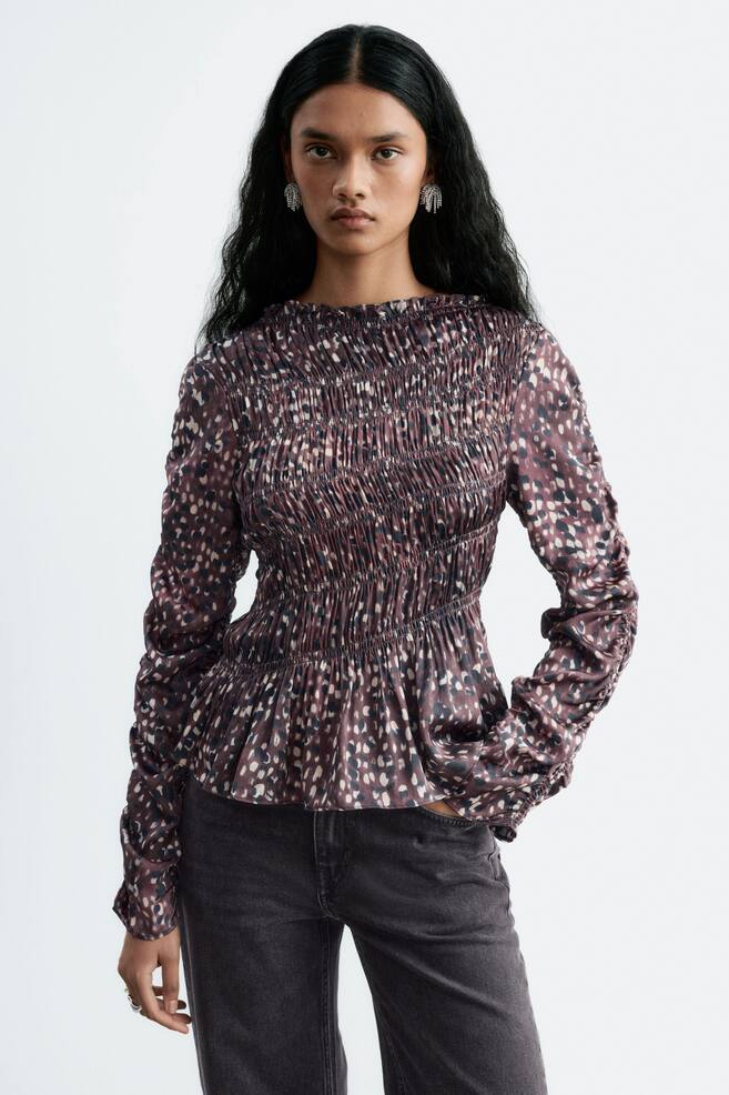 A smocked blouse featuring a high, ruffled neckline, long sleeves, and an abstract pattern is being worn by the model. The blouse has a fitted, smocked bodice and is paired with dark grey jeans. - 1