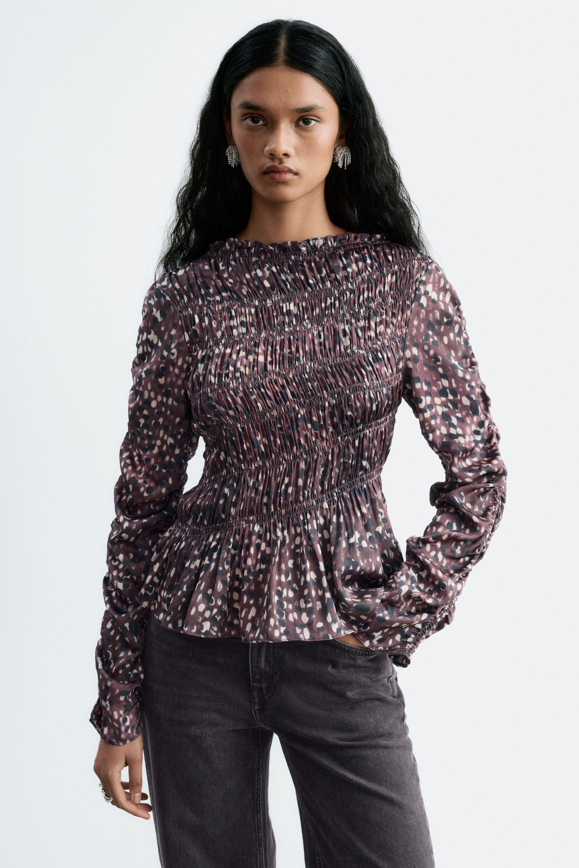 View larger image: A smocked blouse featuring a high, ruffled neckline, long sleeves, and an abstract pattern is being worn by the model. The blouse has a fitted, smocked bodice and is paired with dark grey jeans.