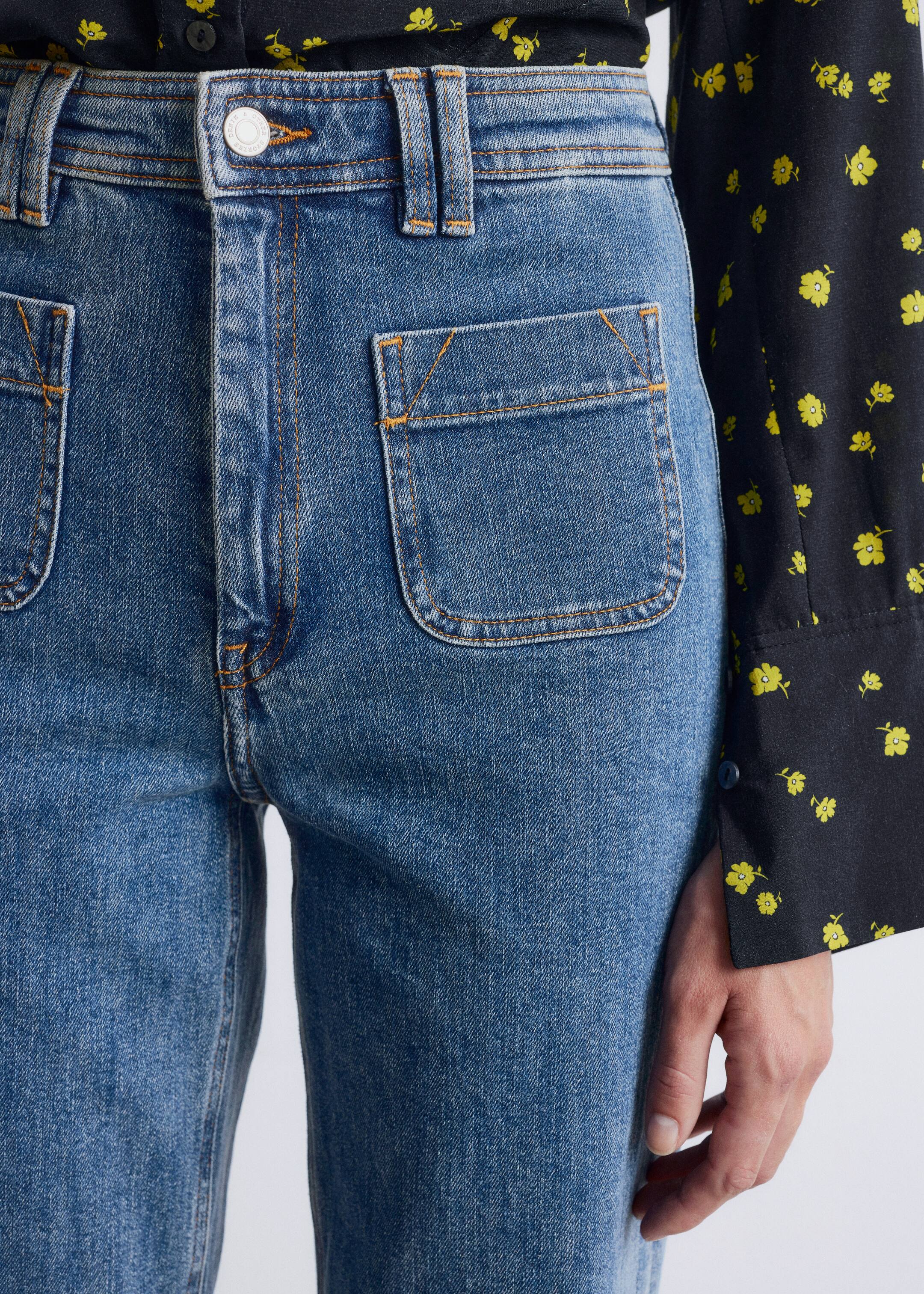 Grotere afbeelding bekijken: Blauwe spijkerbroek wordt van voren getoond, iets schuin naar rechts, met een medium wassing, opvallende oranje contraststiksels, grote afgeronde steekzakken op de voorkant van de heupen en een zilverkleurige knoop die de tailleband sluit. Een zwart bloemenshirt wordt in de jeans gestopt.