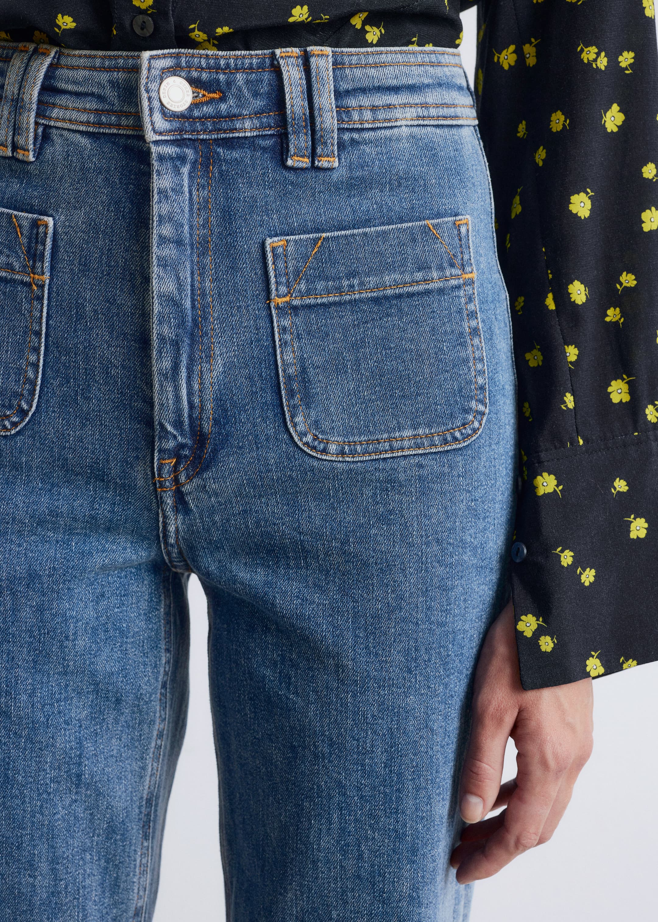Größeres Bild anzeigen: Blaue Denim-Jeans werden von vorne gezeigt, leicht nach rechts geneigt, mit einer mittleren Waschung, auffälligen orangefarbenen Kontrastnähten, großen abgerundeten Aufsatztaschen an den vorderen Hüften und einem silberfarbenen Knopf, der den Bund schließt. Ein schwarzes Blumenhemd wird in die Jeans gesteckt.