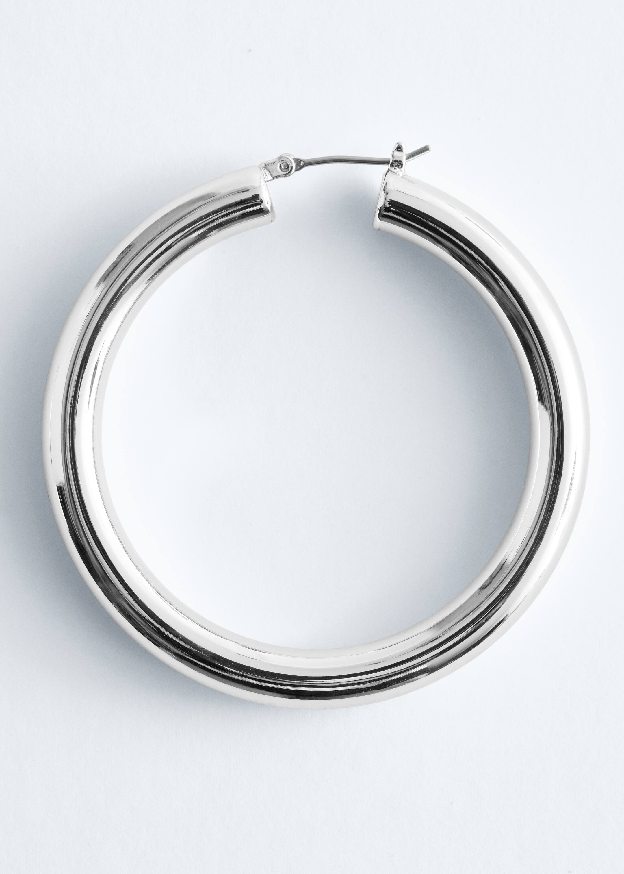 Visa större bild: En stor, silvrig hoop-örhänge presenteras som FRAMSIDAN, med en tjock, rundad tubformad design. Ett gångjärnspostlås är synligt upptill.