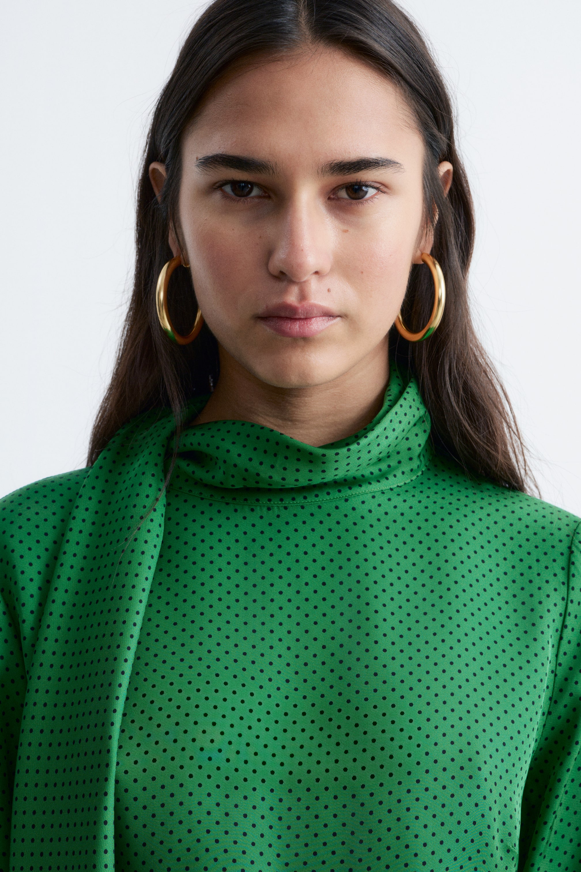 Se større billede: En grøn top med små sorte polkaprikker har en høj, draperet halsudskæring. Et bredt stofpanel strækker sig fra venstre side af halsudskæringen og draperer ned foran.