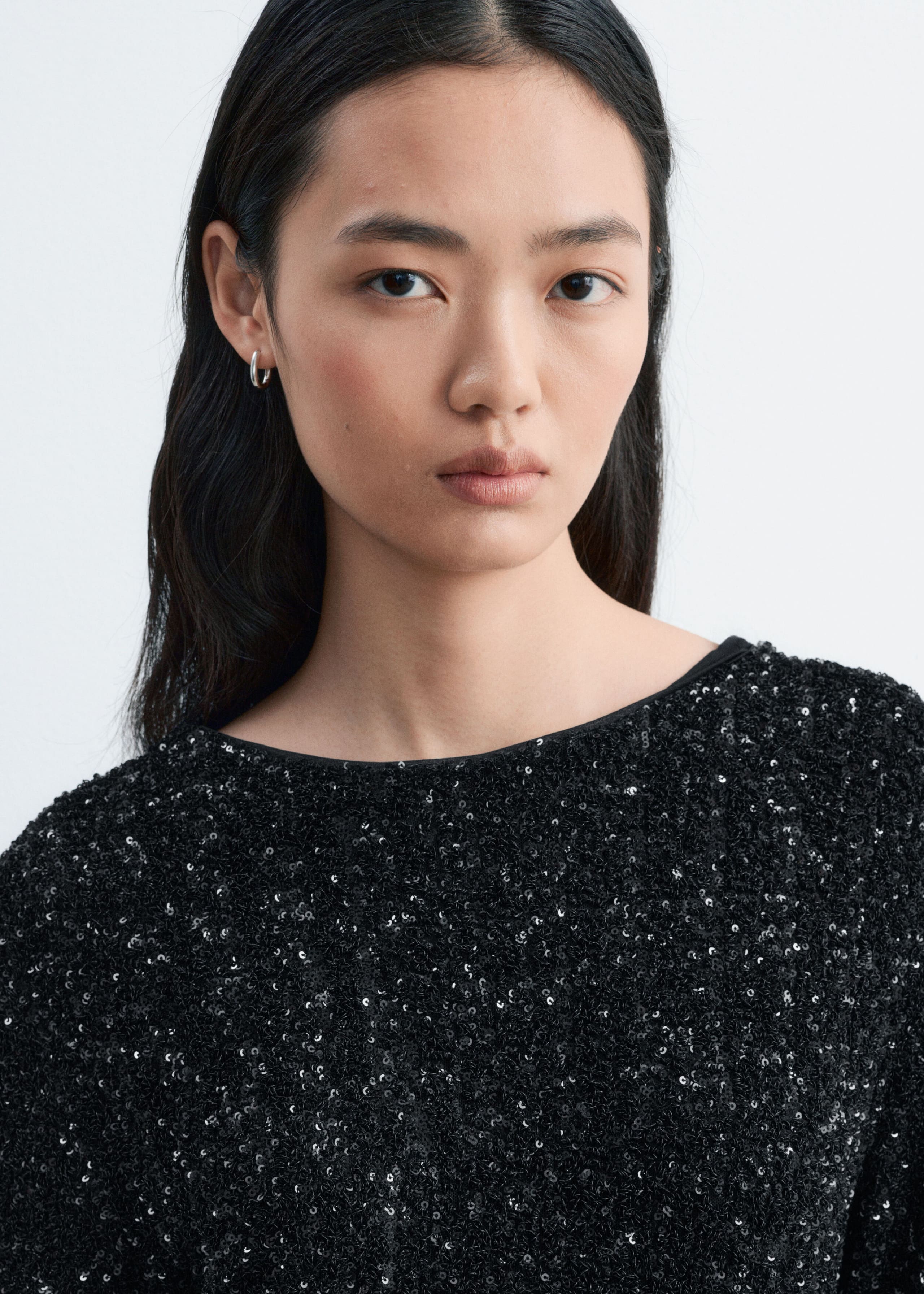 A black sequined dress with a wide neckline is being worn by the model. The garment is covered in small, shimmering black sequins that create a textured surface.