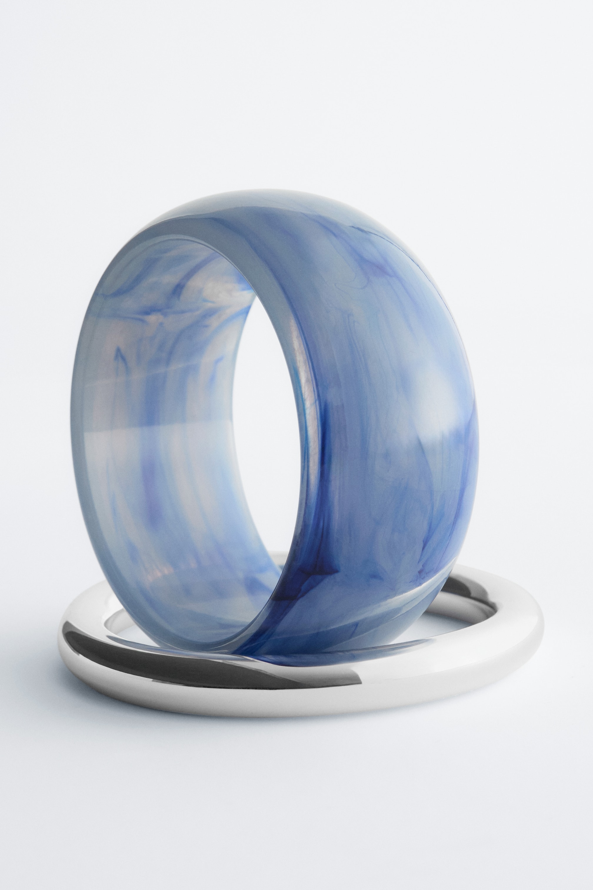 Ver imagen más grande: El conjunto de brazaletes muestra un brazalete azul translúcido de gran tamaño con un patrón marmoleado, colocado en posición vertical y descansando sobre un brazalete de metal plateado pulido y elegante. El brazalete azul presenta su lado frontal, exhibiendo su perfil grueso y redondeado.