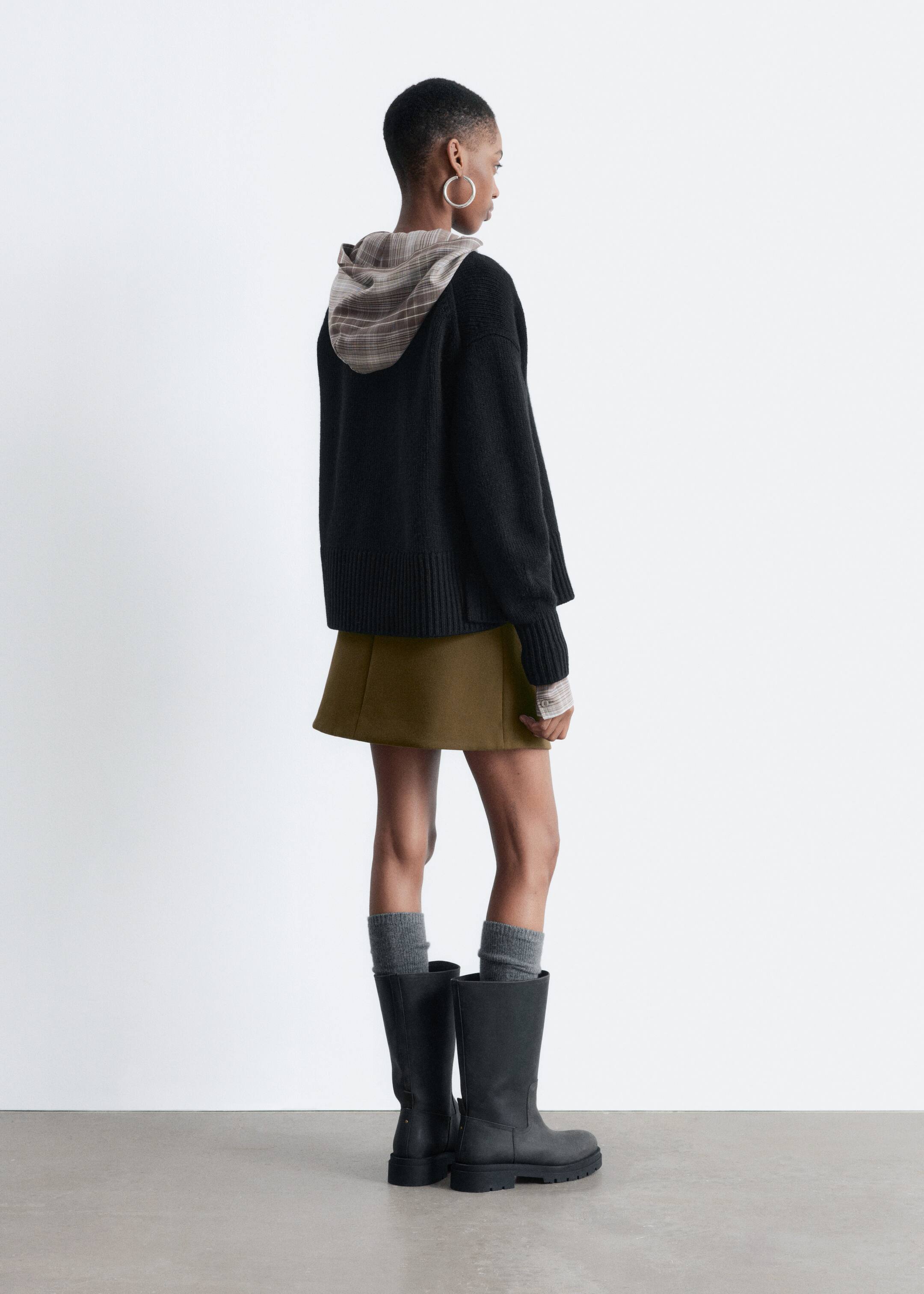 Agrandir l'image: Un cardigan noir en tricot, avec une silhouette ample et des bords côtes, est porté par le mannequin, superposé à un haut à capuche à carreaux.