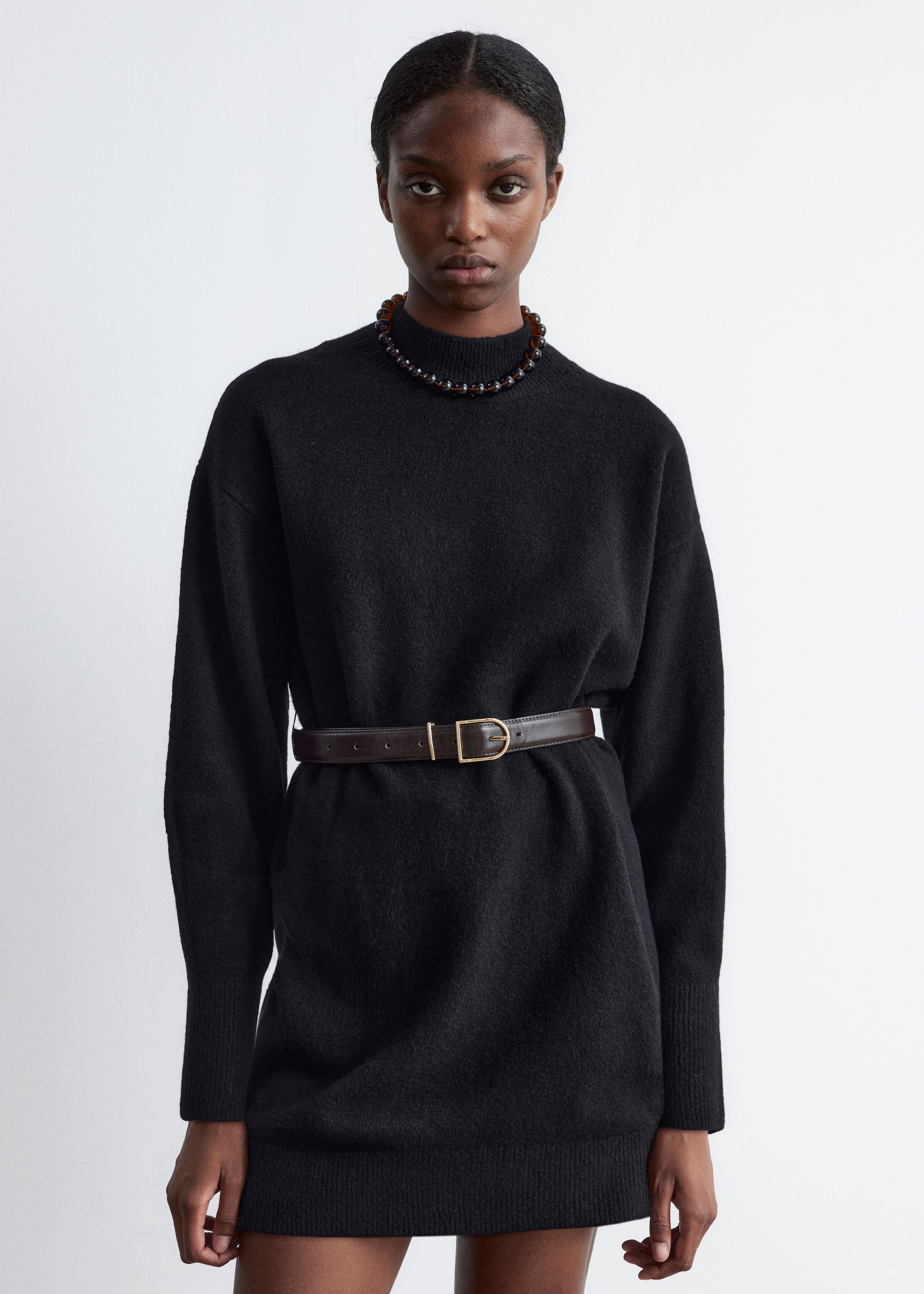 Größeres Bild anzeigen: Ein schwarzes Strick-Minikleid mit Stehkragen und langen Ärmeln wird von dem Model getragen. Ein brauner Gürtel mit einer goldfarbenen Schnalle schnürt das Kleid an der Taille, und eine Perlenkette wird über dem Stehkragen getragen.