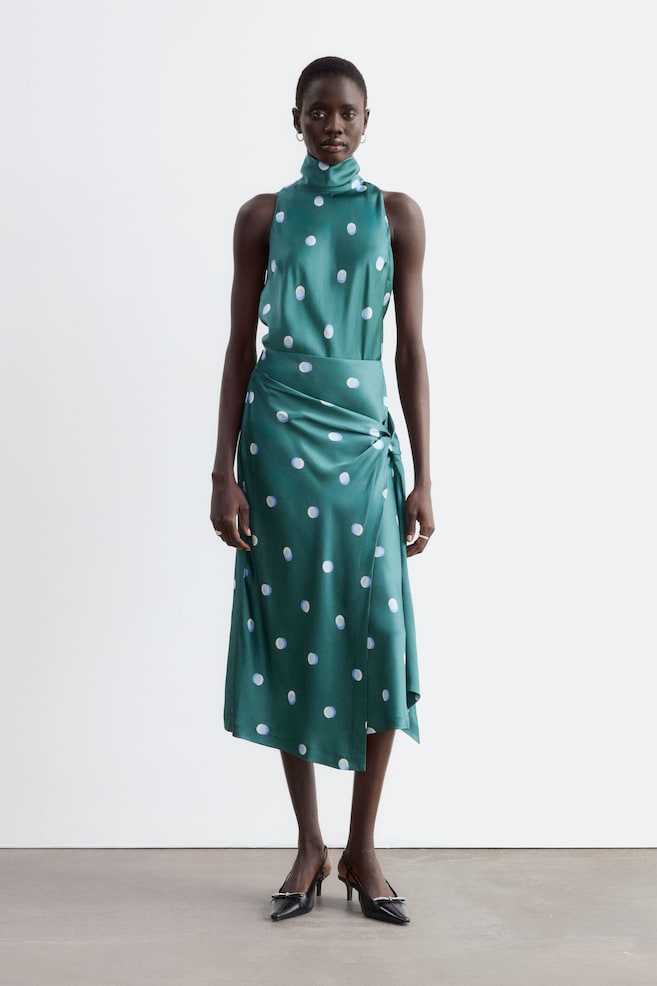 A green midi skirt with a white and blue polka dot pattern is being worn by the model, featuring a draped knot detail at the hip and an asymmetric hem. The skirt is paired with a matching sleeveless high-neck top and black pointed-toe slingback heels. - 1