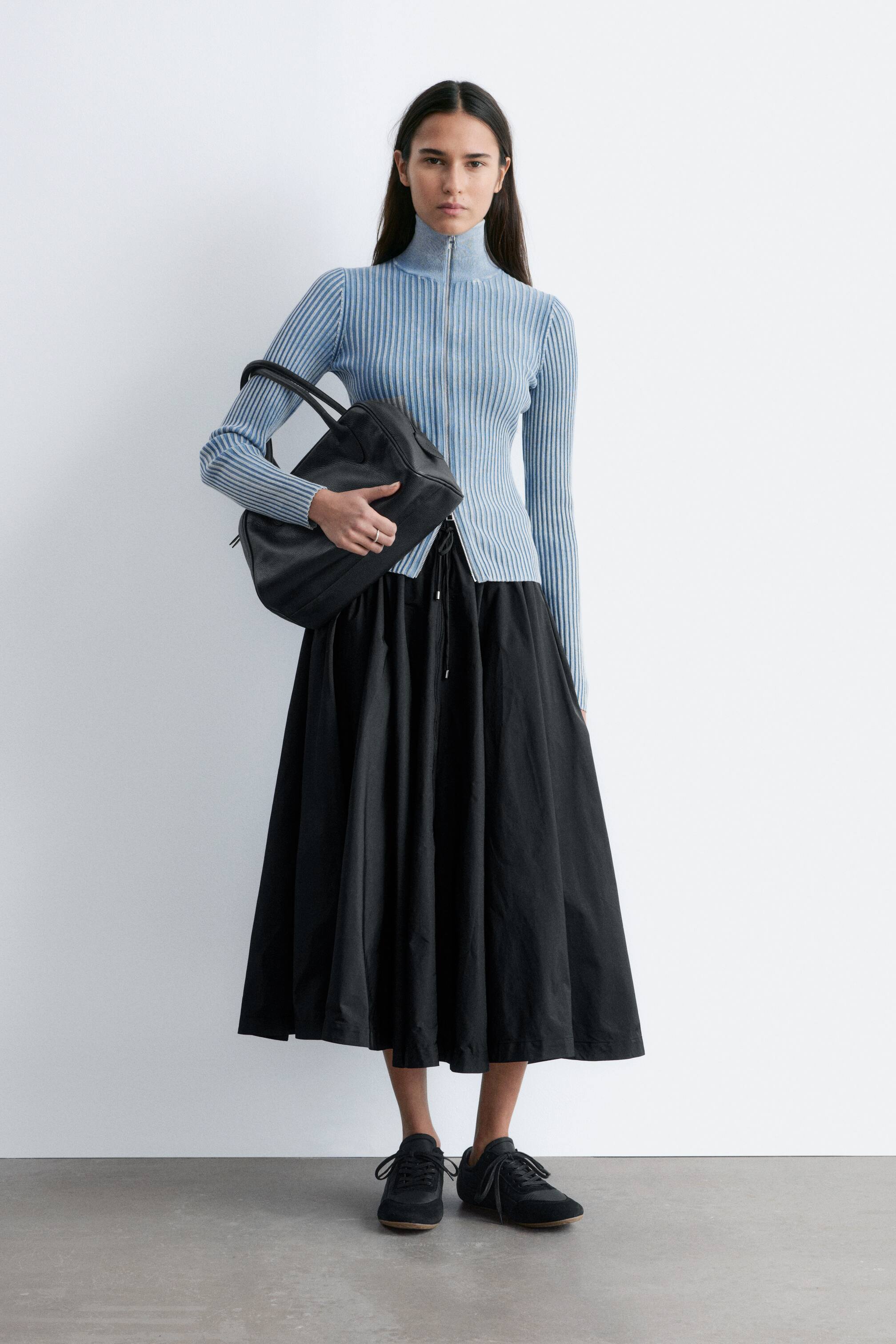 View larger image: The light blue ribbed knit top is displayed from a front view, featuring a high mock neck and a dark-toned zip running down the centre front. This long-sleeved top showcases a vertical ribbed texture and a form-fitting silhouette.