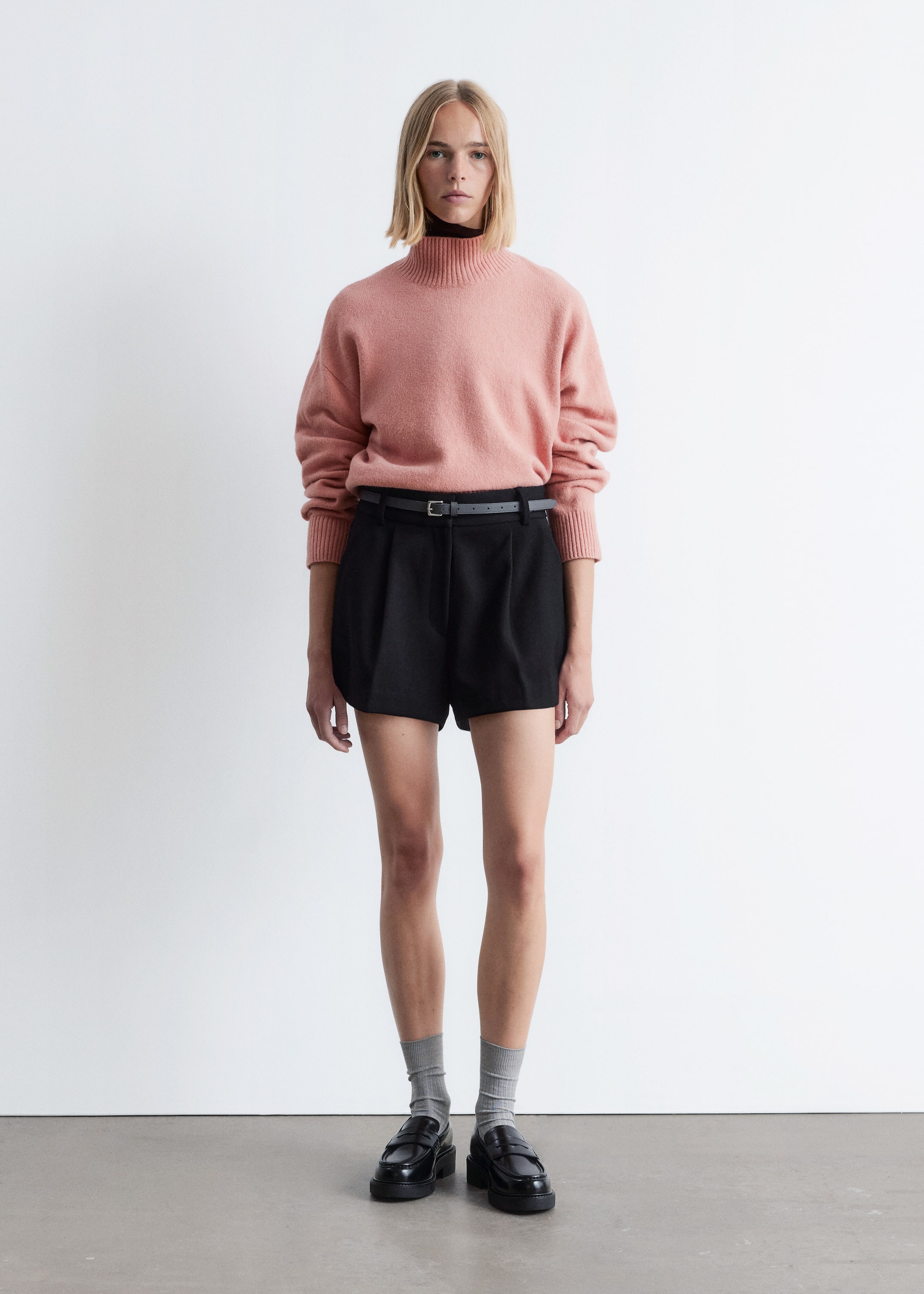 Größeres Bild anzeigen: Ein hellrosa Mock-Neck-Pullover mit entspannter Passform und gerippten Bündchen wird über einem schwarzen Rollkragenoberteil getragen. Schwarze, plissierte Shorts, gesichert mit einem dünnen grauen Gürtel, werden mit schwarzen Loafern mit dicker Sohle und grauen, gerippten Socken gestylt.